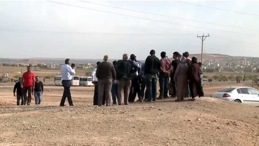 L'image montre un groupe de personnes rassemblées sur un terrain semi-aride. Ils semblent observer quelque chose au loin, peut-être un événement ou une scène particulière. L'arrière-plan est dégagé, avec des collines et des bâtiments visibles au loin. Il y a des véhicules stationnés, dont une voiture à droite. L'atmosphère suggère une interaction entre les gens, qui semblent concentrés sur ce qu'ils regardent. Des câbles électriques sont visibles, ajoutant à l'ambiance rurale de la scène.