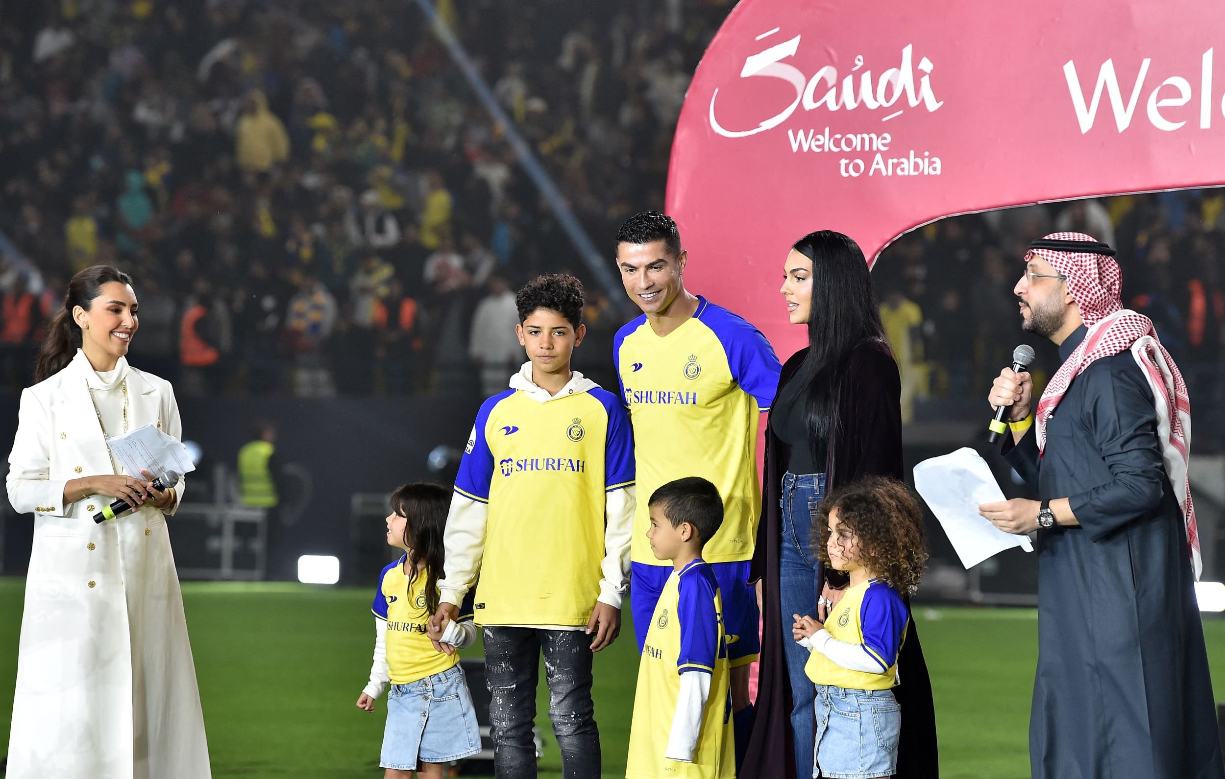 L'image montre une scène sur un terrain de football, où plusieurs personnes sont réunies autour d'un joueur, probablement un footballeur célèbre, en tenue de jeu. Il y a des enfants, probablement des membres de la famille, qui se tiennent à ses côtés. Une femme, habillée d'une manière élégante, semble s'adresser à l'audience, tandis qu'un homme en tenue traditionnelle parle également en tenant un microphone. En arrière-plan, des spectateurs sont visibles, et une grande arche avec les mots "Welcome to Arabia" est installée, indiquant un événement spécial.