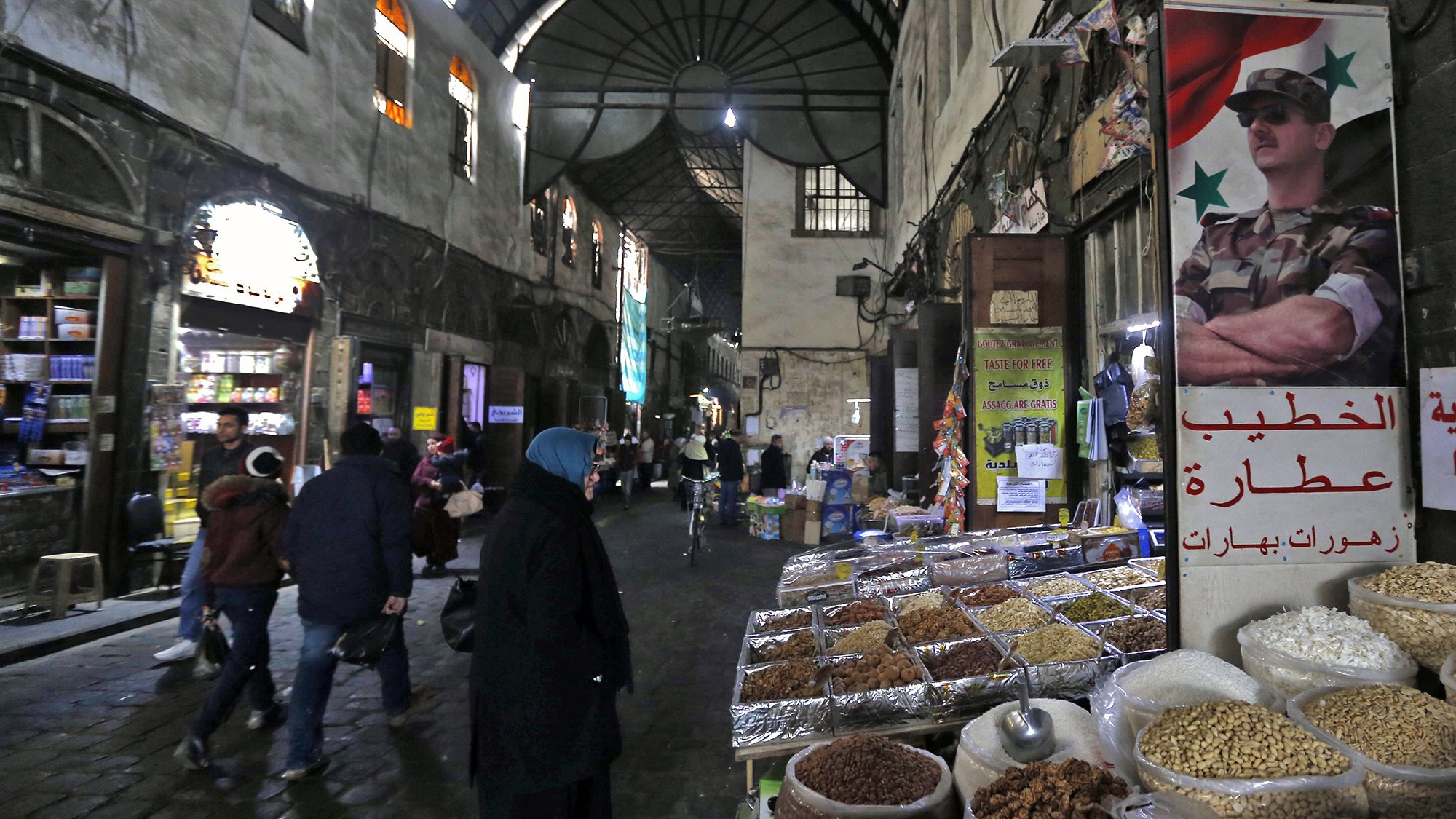 L'image montre une scène d'un souk ou d'un marché typique, probablement en Syrie. On aperçoit des étals avec des épices, des fruits secs, et d'autres produits en vente. Les murs de la rue sont en pierre et semblent anciens, créant une ambiance historique. Des personnes, dont certaines portent des vêtements traditionnels, se déplacent dans la rue, tandis qu'une affiche avec un symbole militaire est accrochée à un mur, ajoutant un élément de contexte culturel et politique à la scène. La lumière faible et les ombres accentuent l'ambiance conviviale et animée du marché.
