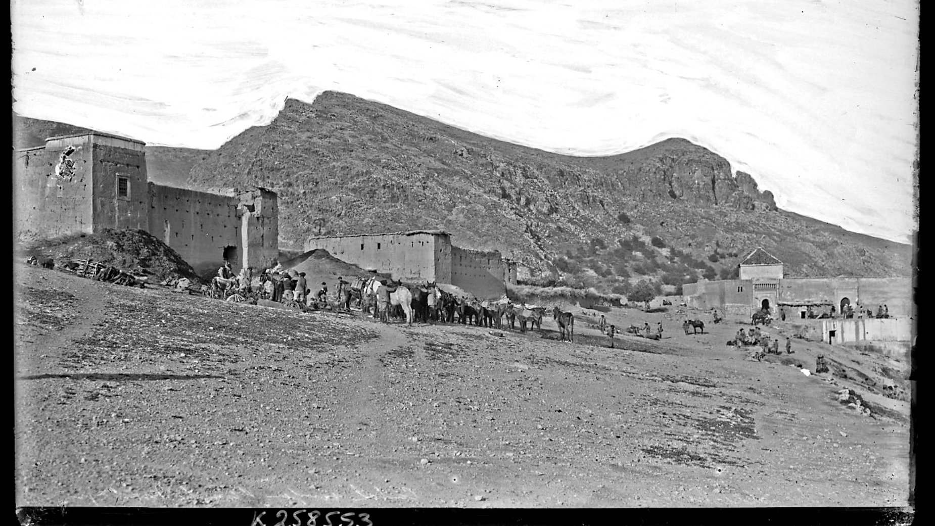 L'image présente un paysage désertique avec des montagnes en arrière-plan. À gauche, on aperçoit des bâtiments en pierre, probablement des fortifications ou des maisons, qui semblent très anciens. Au premier plan, un groupe de personnes est visible, accompagné de plusieurs animaux de charge, comme des chameaux ou des ânes. Le ciel est clair, montrant une atmosphère paisible, et le terrain est aride, typique des régions montagneuses désertiques. L'ensemble dégage une impression d'histoire et de culture.