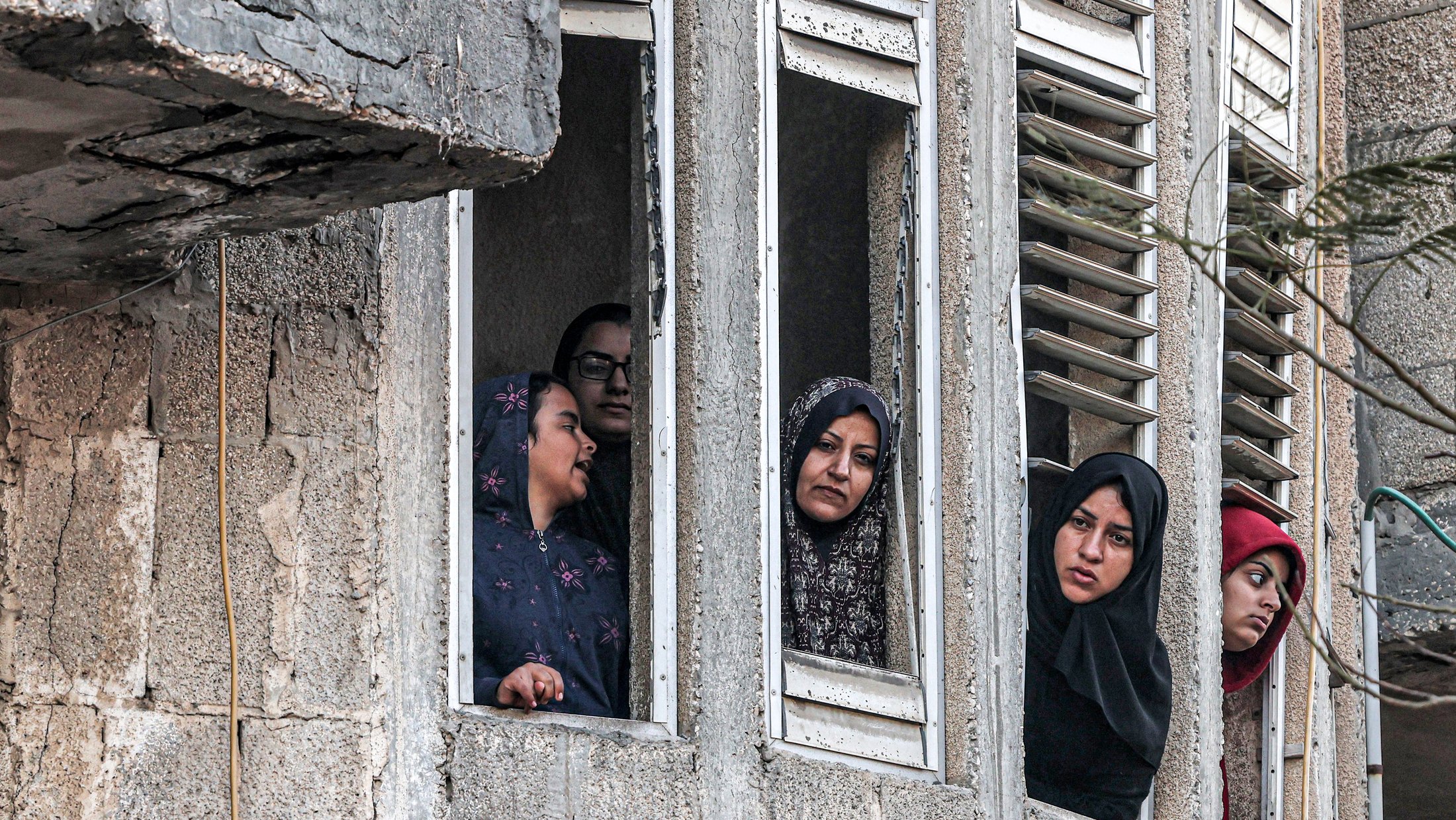 L'image montre plusieurs femmes regardant par des fenêtres d'un bâtiment. Elles semblent attentives et curieuses, certaines portant des hijabs. Le mur extérieur du bâtiment est en béton, et on peut apercevoir des fenêtres aux volets cassés. L'ambiance paraît chargée d'émotions, mais on ne peut pas déterminer le contexte exact de la scène.