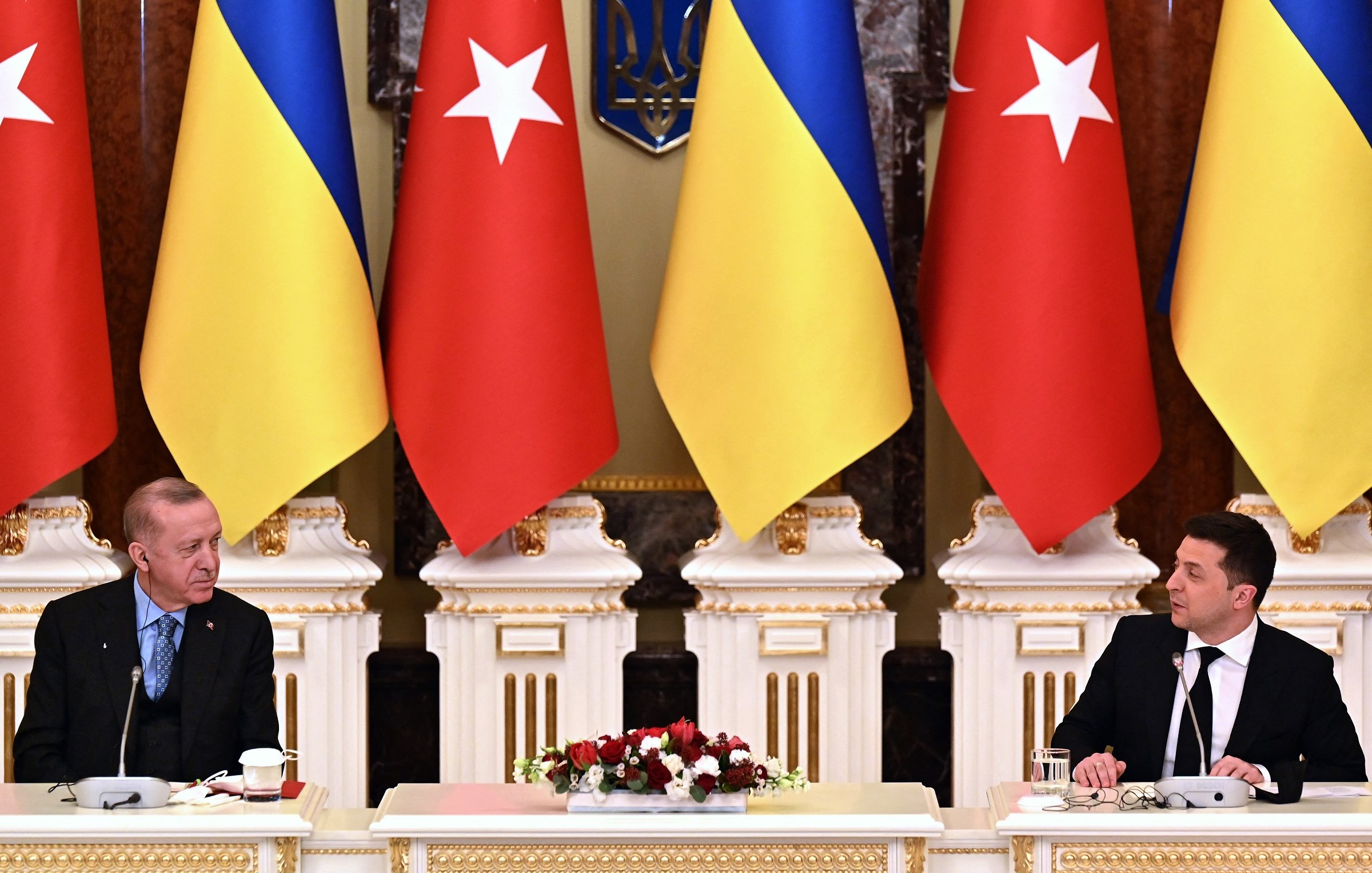 L'image montre une scène officielle avec deux hommes assis à une table. À gauche, un homme porte un costume sombre et semble en discussion. À droite, un autre homme est également en costume, regardant vers son interlocuteur. Derrière eux, on voit des drapeaux turcs et ukrainiens disposés de manière symétrique, et une décoration élégante. L'atmosphère générale évoque une rencontre diplomatique importante.