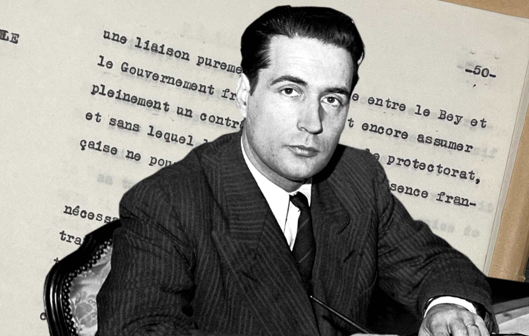 L'image montre un homme en costume assis à une table, tenant un stylo. En arrière-plan, on aperçoit des documents écrits, probablement des notes ou un texte officiel. L'homme a un regard sérieux et semble concentré sur son écrit. Son environnement suggère un contexte historique ou politique.