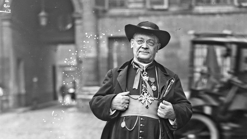 L'image montre un homme en tenue traditionnelle, probablement un ecclésiastique. Il porte une longue robe noire avec des ornements et des boutons sur le devant. Son chapeau est large et il semble poser avec une certaine dignité, les bras légèrement écartés. En arrière-plan, on aperçoit des bâtiments anciens et un véhicule à roues, ce qui suggère une ambiance historique. L'environnement de la rue est pavé, ajoutant à l'atmosphère d'époque.