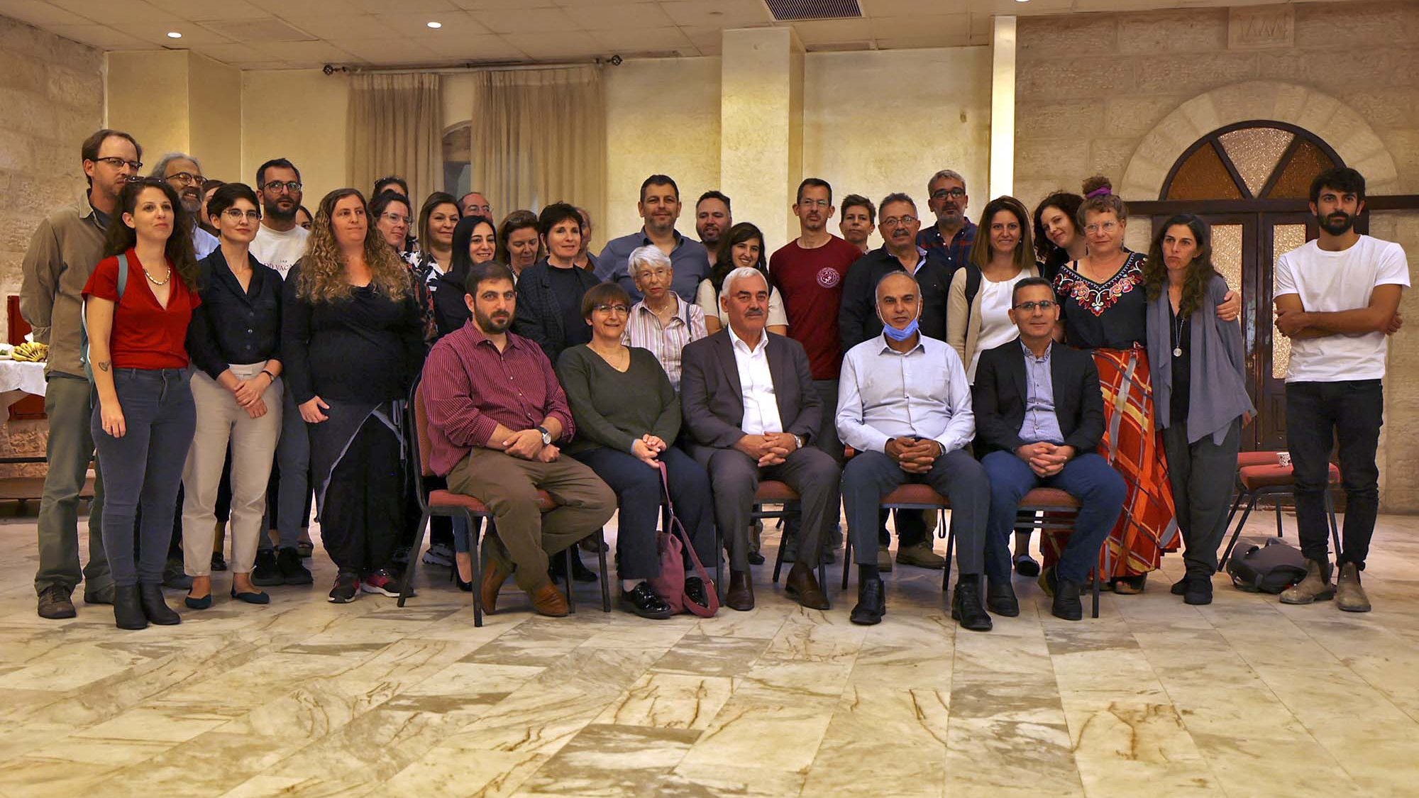 L'image montre un grand groupe de personnes rassemblées dans une salle, probablement pour un événement ou une célébration. Les gens sont assis et debout, souriant et regardant vers l'objectif. L'espace présente des murs en pierre et un sol en marbre, créant une atmosphère conviviale. On peut remarquer une variété de styles vestimentaires parmi les participants, ce qui indique une diversité au sein du groupe.