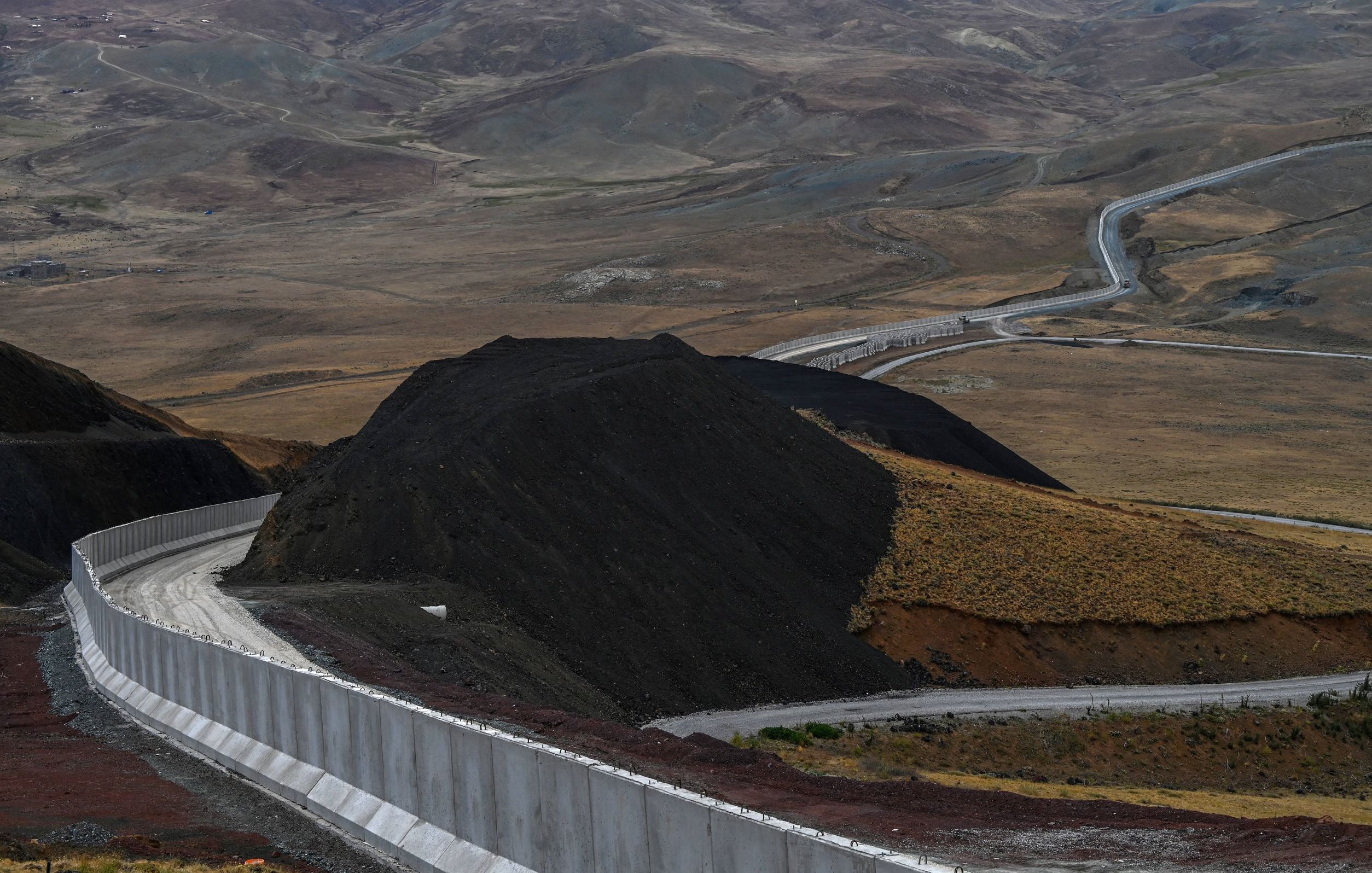 L'image montre un paysage vaste et aride, avec des collines et des montagnes en arrière-plan. Au premier plan, il y a un mur de béton qui serpente le long du sol, suivi d'une route qui s'étend dans la vallée. Le terrain est principalement composé de terre brune et de végétation clairsemée, avec des déclivités et des pentes. L'ambiance générale semble désertique et isolée.