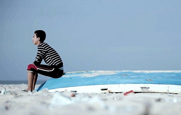 L'image montre un jeune garçon assis sur une coque de bateau échouée sur une plage. Il porte un t-shirt à rayures et un pantalon rouge. L'environnement est clair, avec un ciel bleu pâle et une plage de sable. Le garçon semble pensif, tourné vers l'horizon, tandis que la coque du bateau, abandonnée et usée, ajoute un élément de mélancolie à la scène.