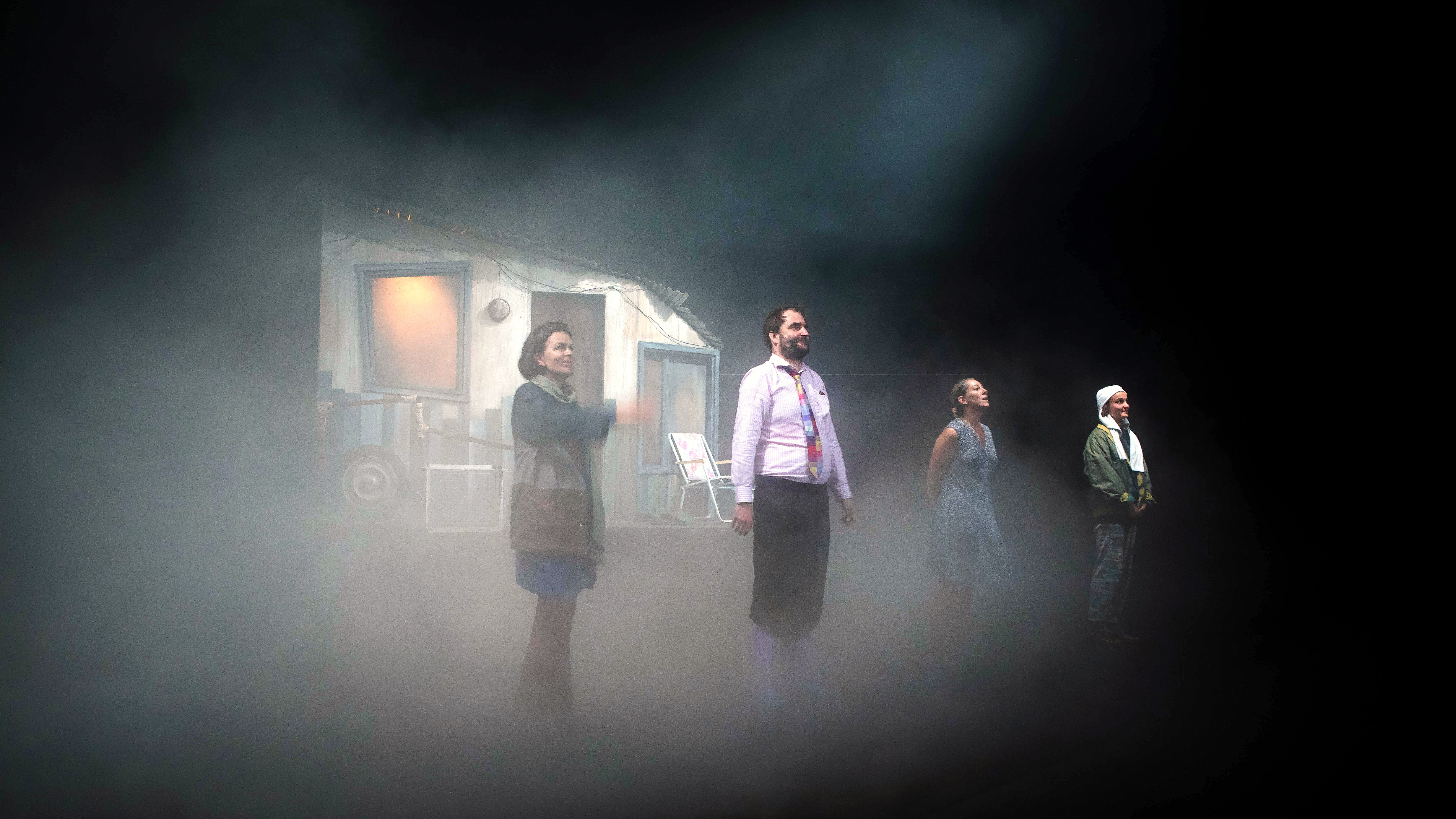 L'image présente une scène de théâtre avec quatre personnages en position debout sur scène. Ils se trouvent dans une atmosphère brumeuse, ce qui ajoute une ambiance mystérieuse. En arrière-plan, on peut apercevoir une petite maison ou un décor qui semble inhabitée. Les personnages sont vêtus de manières diverses, ce qui suggère des rôles ou des traits de caractère différents. La lumière semble tamisée, renforçant l'effet dramatique de la scène.