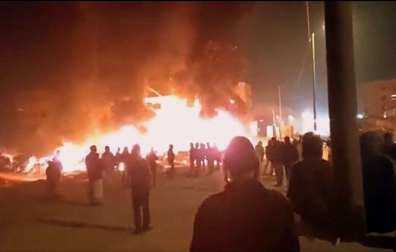 L'image montre une scène nocturne où un bâtiment est enflammé, créant une grande colonne de feu et de fumée. Des personnes se tiennent au premier plan, observant la situation. On peut noter une ambiance de tension, avec des lumières des bâtiments environnants qui ajoutent à l'atmosphère dramatique. Les silhouettes des personnes sont découpées contre la lumière des flammes.