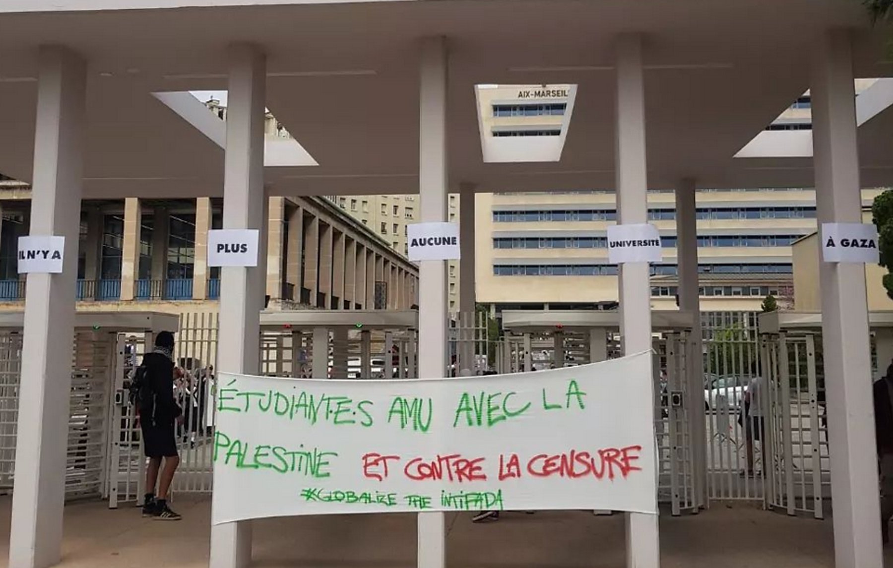 L'image montre l'entrée d'Aix Marseille Université, avec des panneaux affichant des messages. Au premier plan, il y a une grande bannière sur laquelle est écrit en rouge et en vert : "Étudiantes AMU avec la Palestine et contre la censure". L'atmosphère semble refléter un soutien à la cause palestinienne et une dénonciation de la censure. Quelques personnes sont visibles dans la scène, et le cadre est universitaire.