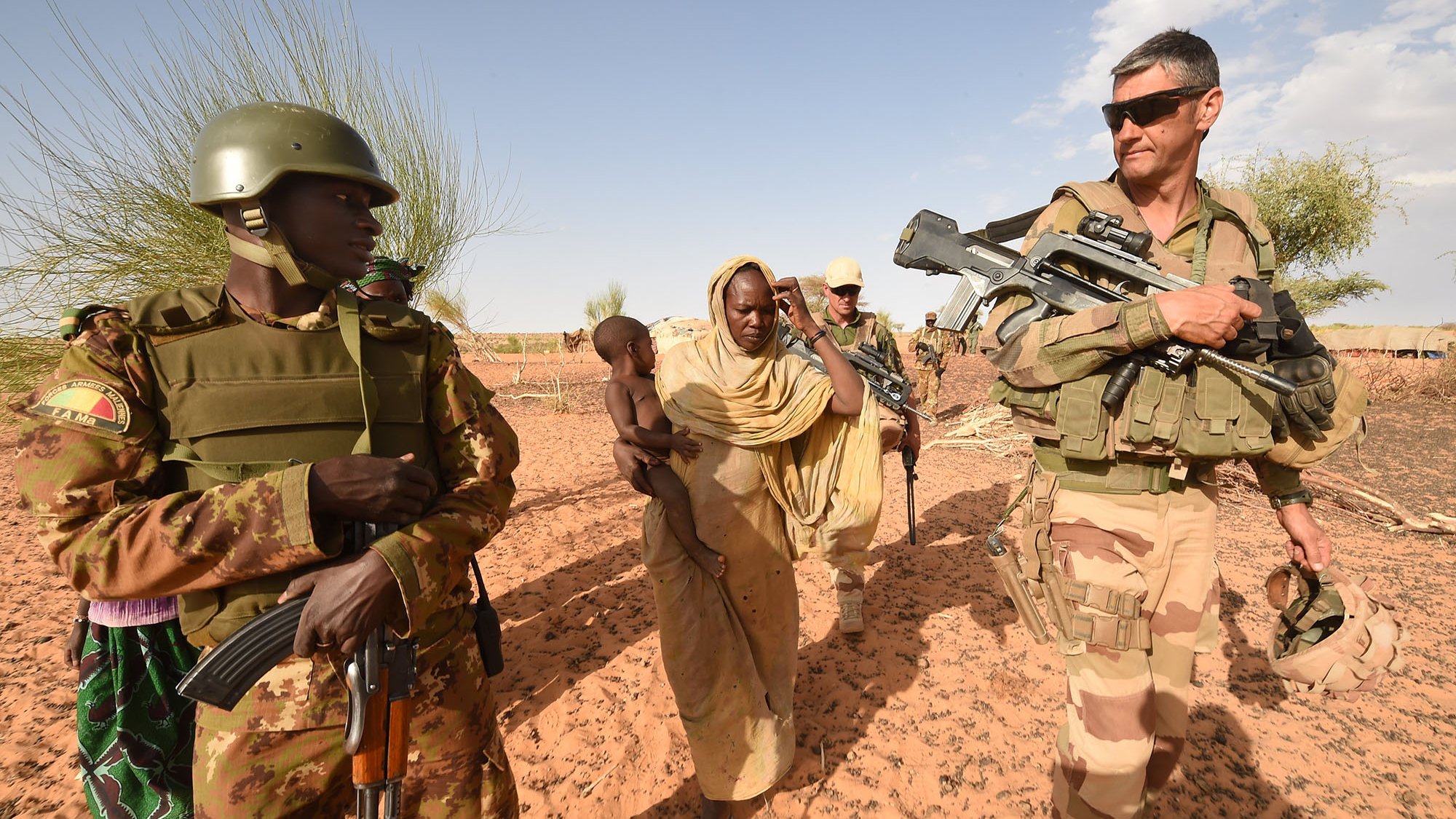 L'image illustre une scène de terrain militaire dans un environnement désertique. On peut voir deux soldats armés, l'un en uniforme de camouflage avec un casque, et l'autre portant un uniforme militaire beige. Ils escortent une femme vêtue d'une robe traditionnelle, qui porte un enfant dans ses bras. L'arrière-plan montre un paysage désertique avec quelques arbres, suggérant une zone aride. Cette image évoque des thèmes de sécurité, de protection et d'interaction humanitaire.