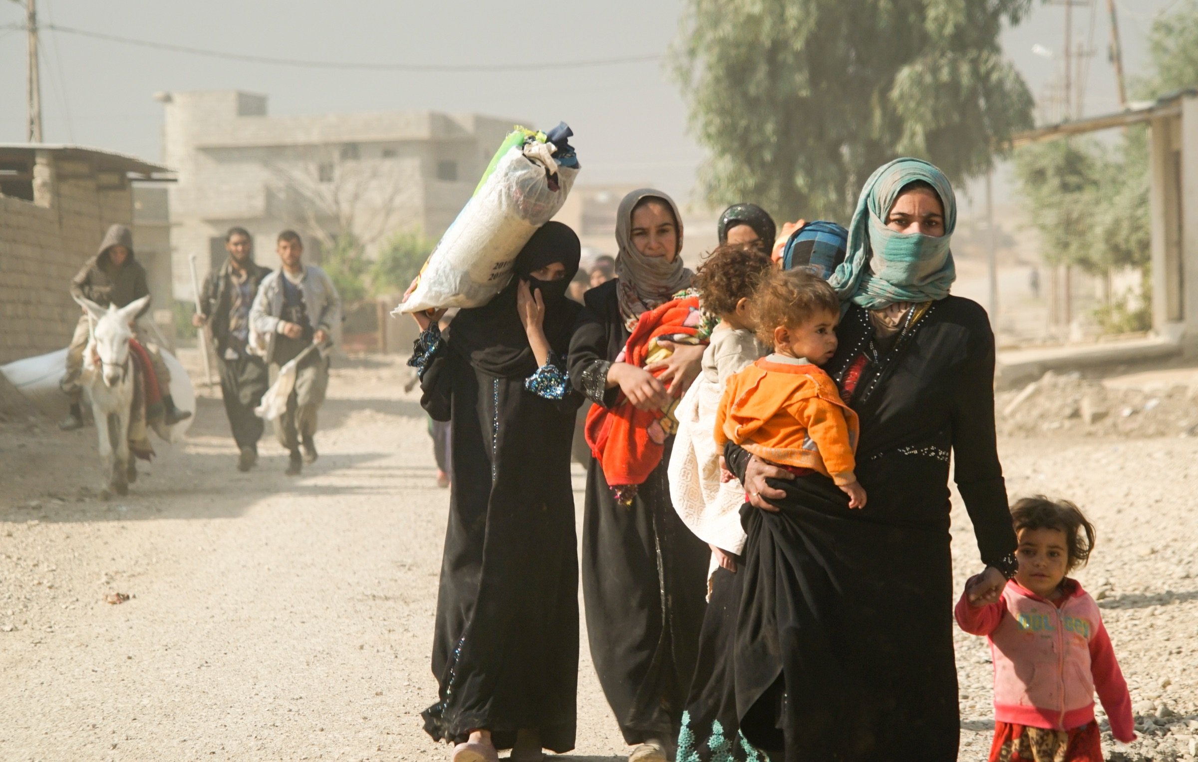L'image montre un groupe de femmes et d'enfants marchant sur un chemin en terre. Certaines femmes portent des vêtements traditionnels et des foulards. L'une d'elles porte un enfant sur son bras, tandis qu'une autre porte un paquet sur sa tête. En arrière-plan, on peut voir des bâtiments et une personne à cheval s'approchant. L'atmosphère semble poussiéreuse et le paysage est rural. Les expressions des femmes sont variées, allant de la concentration à la sollicitude envers les enfants.