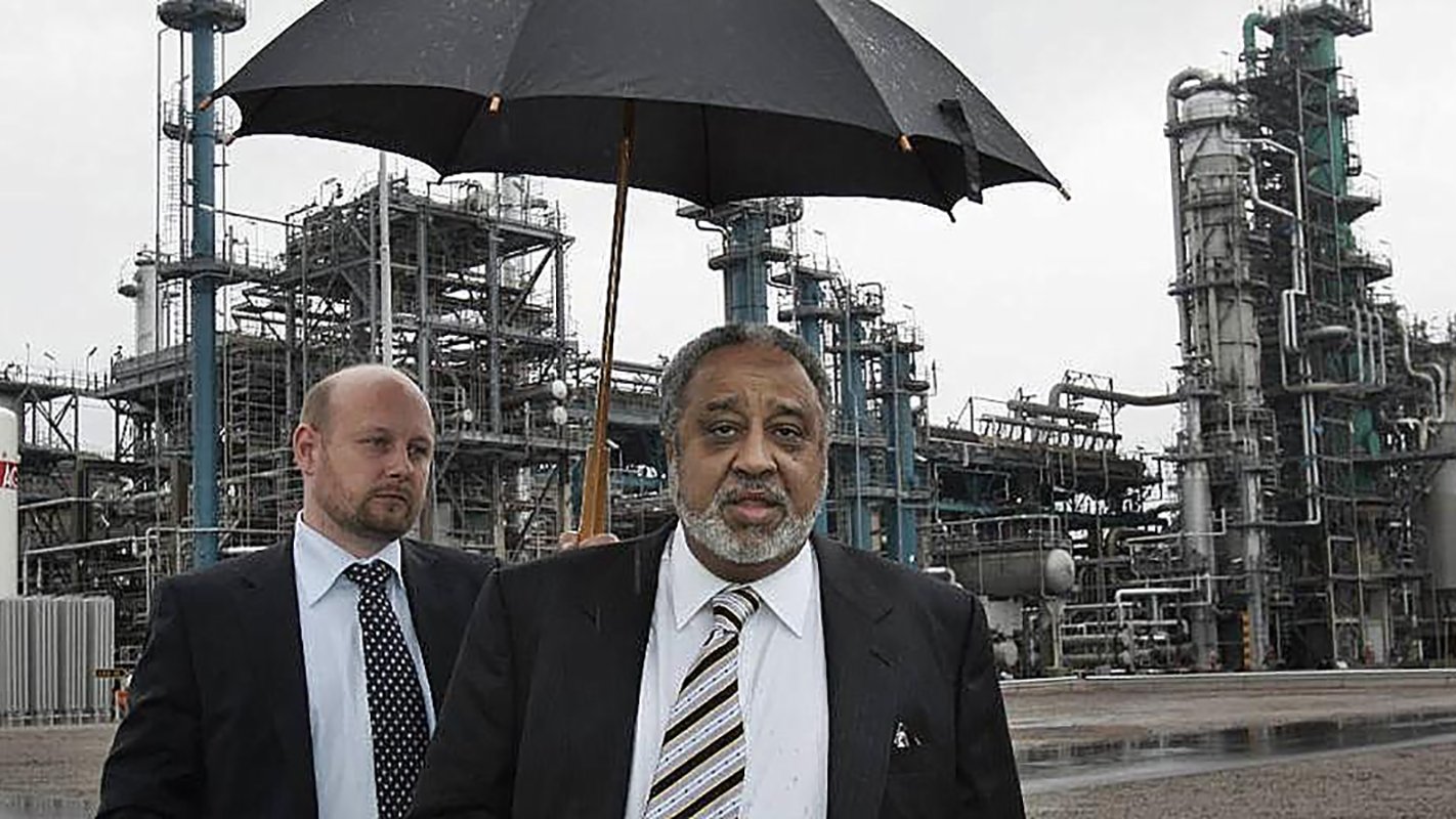 L'image montre deux hommes marchant dans une zone industrielle. L'un des hommes, qui porte un costume noir et une cravate, tient un parapluie, probablement pour se protéger de la pluie. L'autre homme, à l'arrière-plan, est également en costume. On peut voir des structures industrielles, comme des réservoirs et des cheminées, en arrière-plan, sous un ciel grisâtre. L'ambiance générale de l'image est celle d'une visite ou d'une inspection dans un environnement industriel.