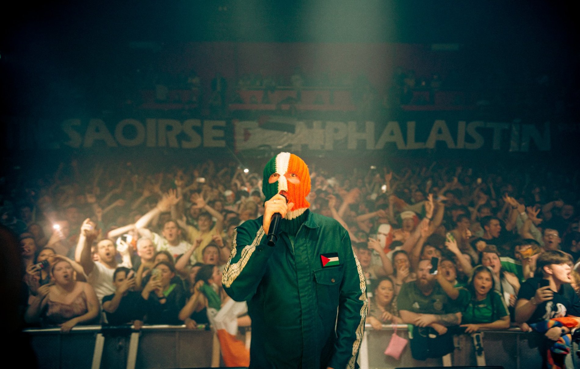 Un artiste en concert, masqué, devant une foule enthousiaste, avec des drapeaux irlandais et palestiniens.