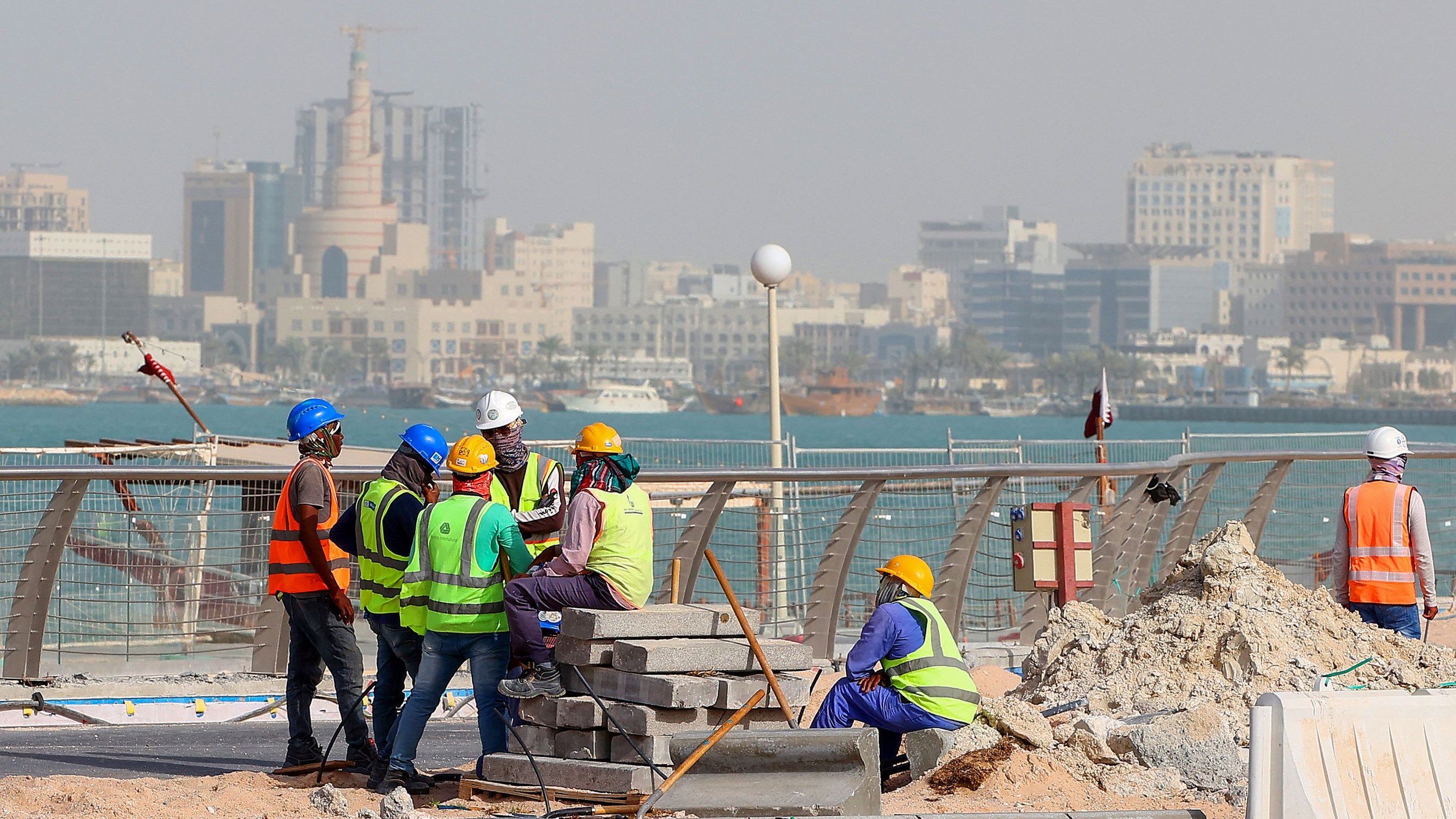 L'image montre un groupe de travailleurs sur un chantier de construction près de la mer. Ils portent des vêtements de sécurité, comme des gilets réfléchissants et des casques. En arrière-plan, on peut voir une ville avec des bâtiments modernes et des structures élevées. L'ambiance semble occupée, et le chantier est environné de matériaux de construction, avec des blocs empilés. La mer est visible au loin, ajoutant un cadre pittoresque à la scène.