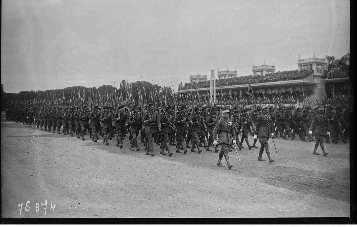 L'image montre un défilé militaire, avec de nombreux soldats marchant en formation. Ils portent des uniformes d'époque et tiennent des fusils à la main. En arrière-plan, on peut voir une tribune avec des spectateurs, probablement des dignitaires ou des spectateurs admirant le défilé. L'atmosphère semble solennelle et organisée, typique des événements militaires de ce genre. Le ciel est nuageux, ce qui donne une ambiance sobre à la scène.