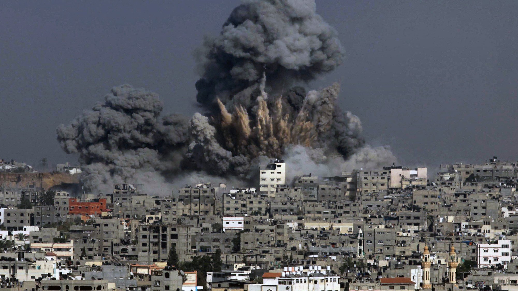 L'image montre une scène de destruction où des colonnes de fumée noire s'élèvent dans le ciel, indiquant qu'une explosion a eu lieu. Les bâtiments sont présents au premier plan, témoignant d'une zone urbaine, et semblent avoir subi des dommages. Le contraste entre la couleur grise de la fumée et les structures environnantes évoque une atmosphère de chaos et de conflit.