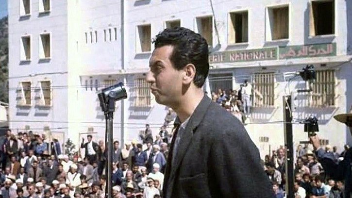 L'image montre un homme se tenant derrière un microphone, semblant s'adresser à une grande foule rassemblée devant lui. L'arrière-plan présente un bâtiment de style architectural simple avec plusieurs fenêtres, possiblement dans un contexte historique ou politique. Les membres de la foule semblent attentifs à son discours. L'éclairage naturel donne une ambiance claire à la scène, suggérant qu'elle pourrait avoir été prise en plein jour.