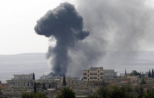 L'image montre un paysage urbain avec un nuage de fumée noir s'élevant dans le ciel, probablement à la suite d'une explosion ou d'un incendie. Les bâtiments aux alentours semblent endommagés, et la végétation est rare. Le fond présente des collines dégradées, ce qui suggère un environnement difficile. L'atmosphère générale évoque une situation de conflit ou de crise.