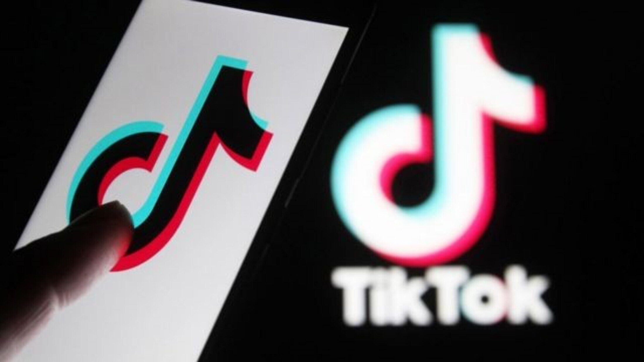 L'image montre un smartphone avec le logo de TikTok affiché à l'écran. On peut voir un doigt en train de toucher ou interagir avec le logo. À l'arrière-plan, il y a le logo de TikTok dans un style similaire, mais peut-être moins détaillé. Les couleurs principales du logo sont le noir, le rouge et le bleu, créant un effet visuel attrayant.