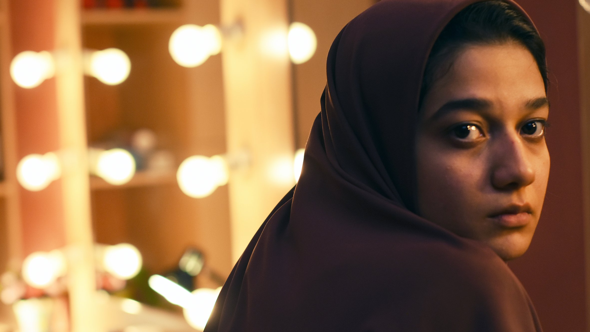 L'image montre une jeune femme avec un regard pensif, portant un hijab. Elle est positionnée de manière à ce que son visage soit tourné légèrement vers la caméra, tandis qu'en arrière-plan, on aperçoit un miroir entouré de lumières douces, créant une ambiance chaleureuse et intime. Son expression reflète une certaine introspection, ajoutant une profondeur émotionnelle à la scène.