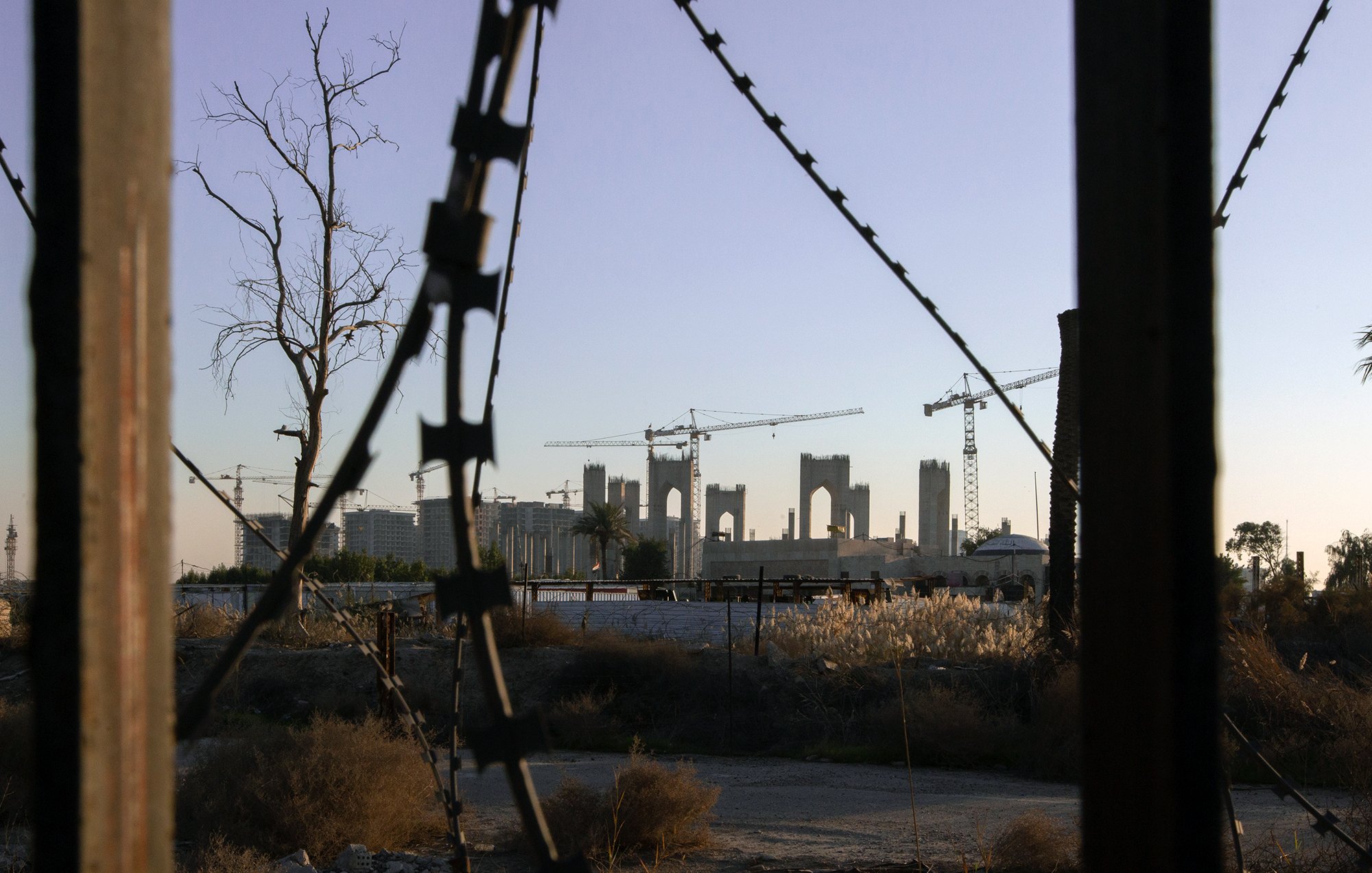 L'image présente un paysage urbain en transformation, avec des structures inachevées à l'arrière-plan, entourées de grues de construction. En premier plan, on peut voir des barbelés et des éléments de végétation desséchée, suggérant un environnement abandonné ou en déclin. La lumière douce du crépuscule ajoute une ambiance mélancolique à la scène, mettant en contraste l'espoir de la construction avec la désolation du terrain environnant.