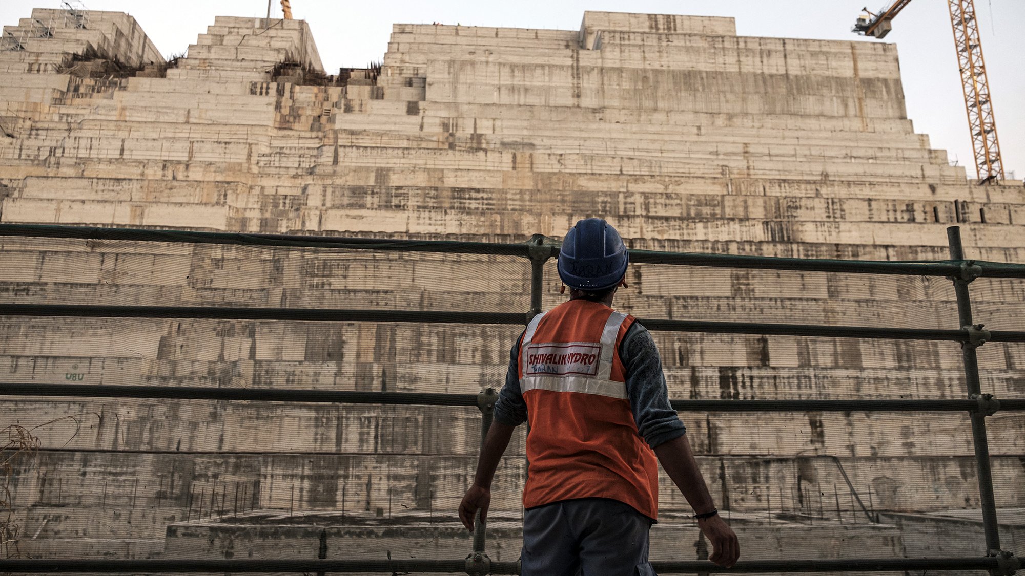 L'image montre un ouvrier sur un chantier de construction, portant un gilet de sécurité orange et un casque. Il se tient devant une grande structure en béton, apparemment inachevée, qui est composée de plusieurs niveaux superposés. En arrière-plan, on aperçoit une grue, ce qui indique que le site est actif. L'atmosphère semble à la fois industrielle et impressionnante, mettant en avant l'ampleur du projet de construction.
