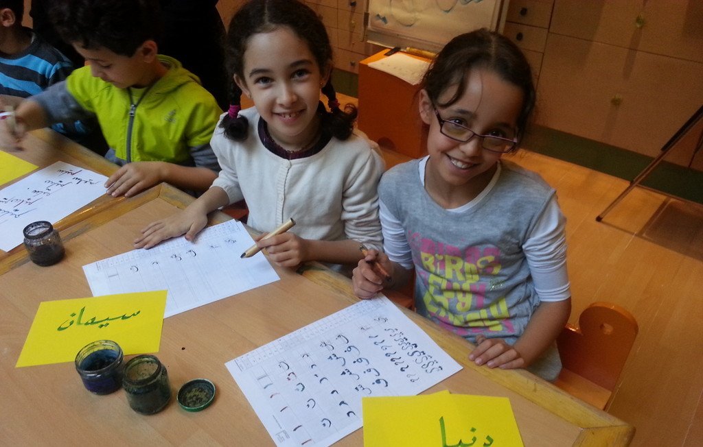 L'image montre deux jeunes filles assises à une table, souriant et travaillant sur des feuilles de papier. Elles utilisent des pinceaux et de l'encre, et semblent engagées dans une activité créative, probablement de calligraphie. Des étiquettes jaunes sont visibles, sur l'une est écrit "سيماء" et sur l'autre "دنيا" en arabe. L'environnement est scolaire, avec d'autres enfants visibles en arrière-plan.