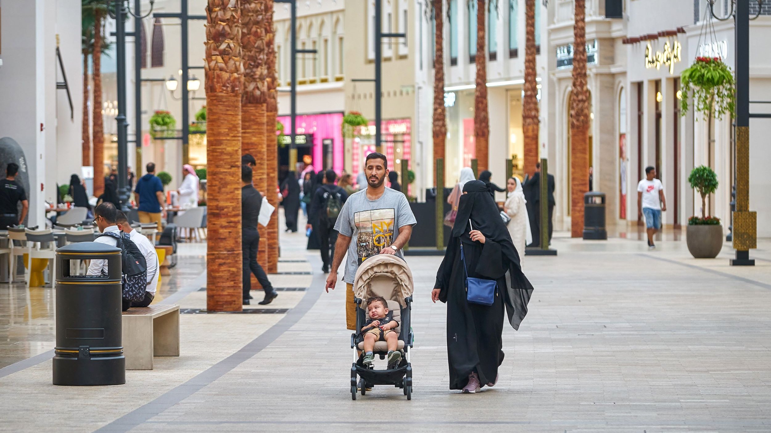 L'image montre une scène animée dans un centre commercial. On peut voir des palmés en arrière-plan et des magasins de différentes boutiques. Un homme marche avec un enfant dans une poussette, tandis qu'une femme, vêtue d'une abaya, les accompagne. Plusieurs autres personnes sont visibles dans l'allée, créant une ambiance de vie urbaine dynamique. La lumière est claire et l'atmosphère semble accueillante.