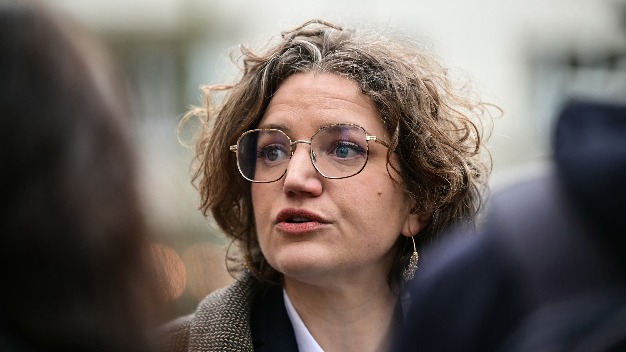 L'image montre une femme avec des cheveux bouclés et des lunettes, en train de parler. Elle semble engagée dans une conversation, peut-être lors d'un événement public. Son expression faciale est sérieuse, et elle porte un vêtement avec un col blanc et une veste. L'arrière-plan paraît flou, suggérant qu'elle se trouve au milieu d'un groupe ou d'une foule.