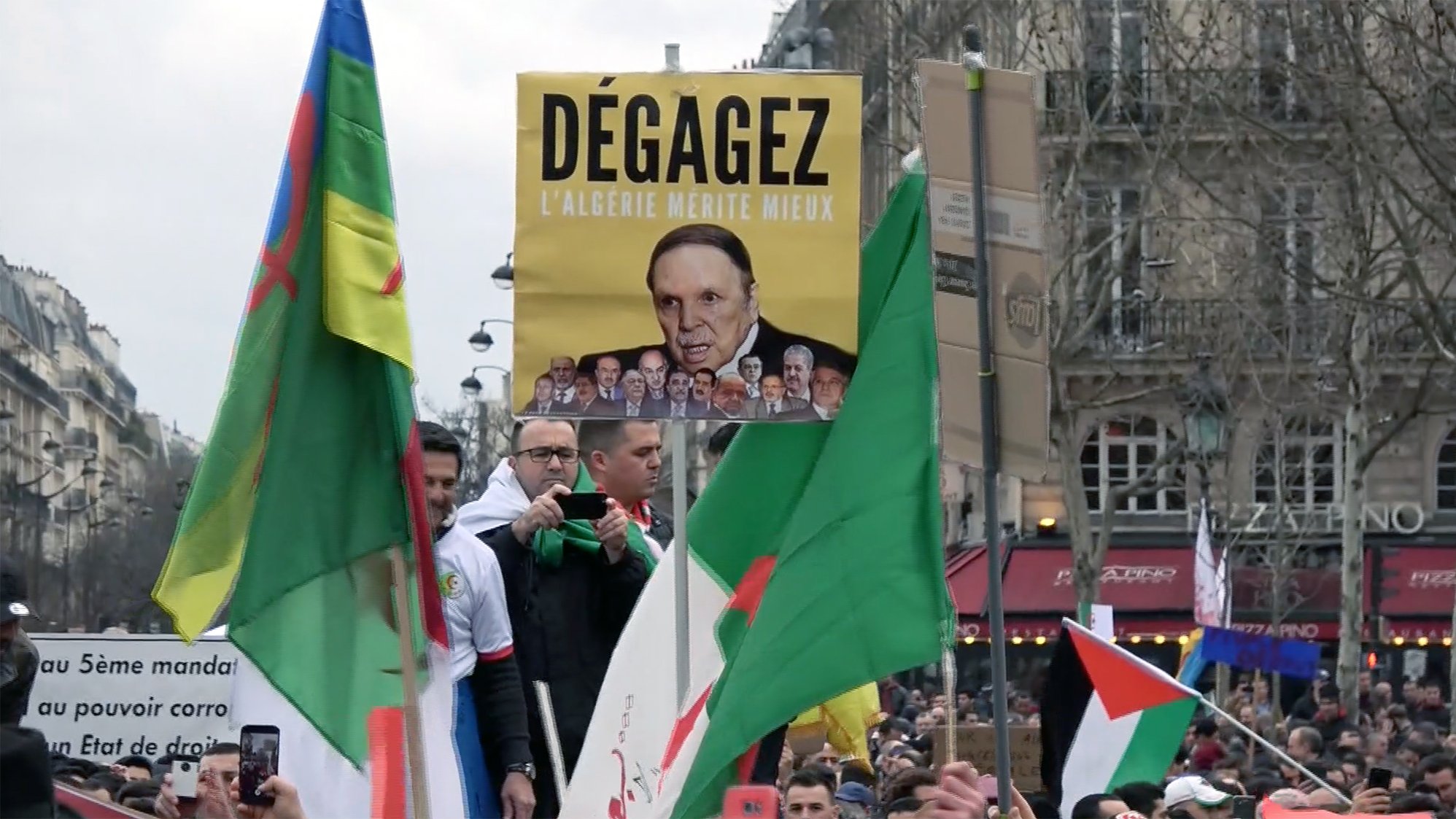 L'image montre une manifestation avec des participants portant des drapeaux. Au centre, un homme tient une grande pancarte sur laquelle est écrit "Dégagez l'Algérie mérite mieux", accompagnée d'une photo d'une personne. Les manifestants semblent exprimer des opinions politiques, et l'atmosphère semble être celle d'une contestation pacifique. Des slogans sur les pancartes reflètent des revendications pour des changements en Algérie.
