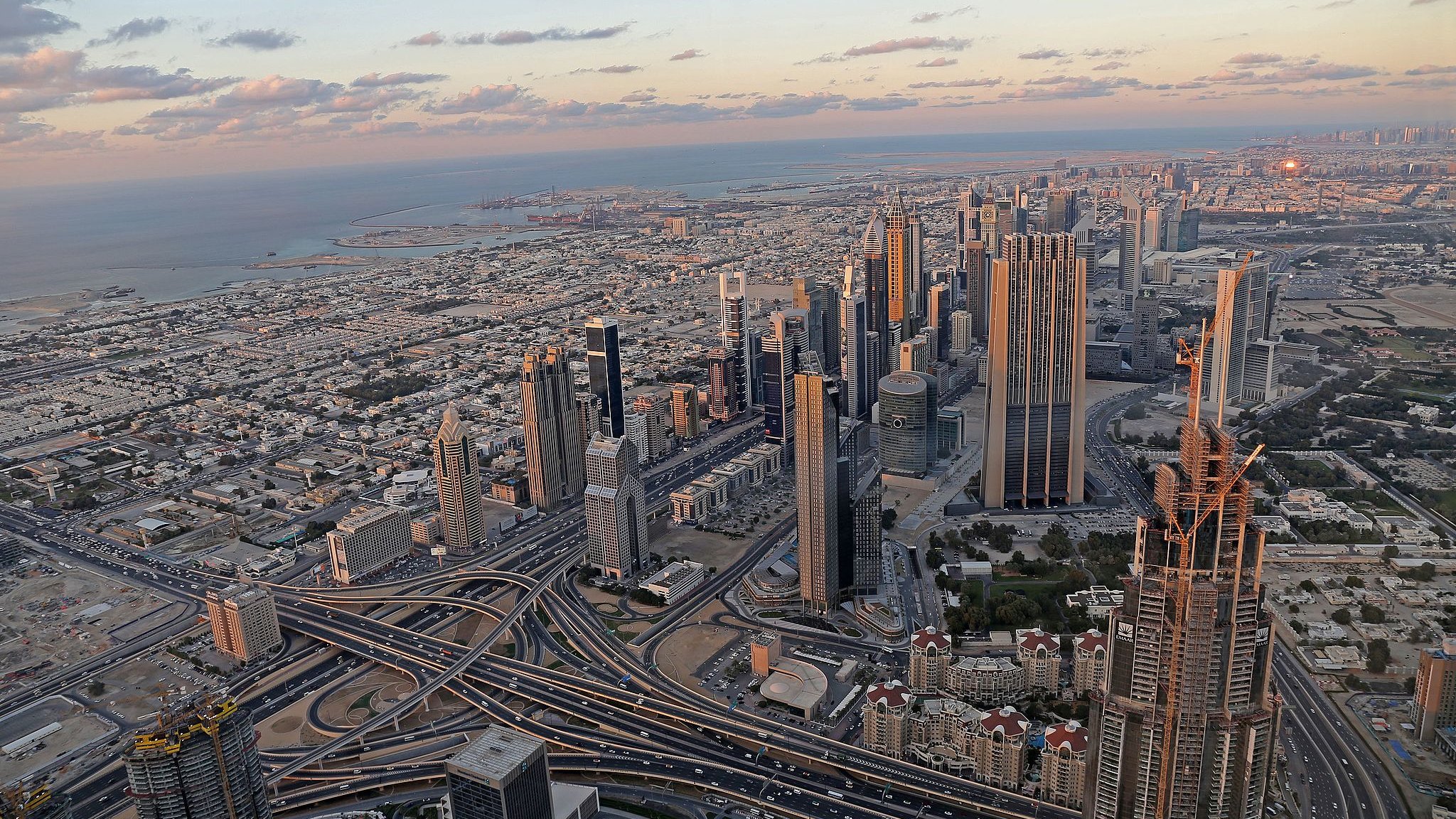 L'image montre un panorama impressionnant d'une ville moderne, probablement Dubai. On y voit de nombreux gratte-ciels élégants s'élevant vers le ciel, ainsi qu'un réseau complexe de routes et d'autoroutes serpentant à travers la métropole. À l'arrière-plan, on aperçoit la mer et des paysages urbains qui s'étendent à perte de vue. Le ciel présente des teintes douces de bleu et d'orange, témoignant d'un coucher de soleil, ce qui ajoute une ambiance tranquille à la scène urbaine animée.