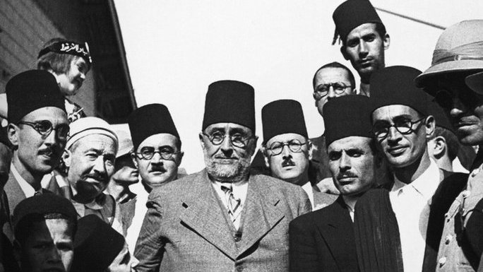 L'image représente un groupe d'hommes en costumes formels, majoritairement portant des fez. Ils semblent être engagés dans une discussion ou un moment de camaraderie. L'ambiance est sérieuse, et les expressions des visages montrent une concentration ou une détermination. Une femme est également visible en arrière-plan, portant un foulard. Le cadre pourrait indiquer un événement historique ou un rassemblement important, probablement dans un contexte culturel ou politique.