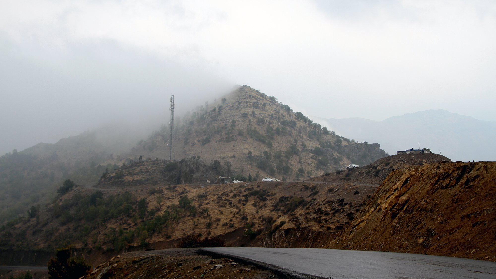 L'image montre un paysage montagneux avec des collines voilées par le brouillard. En arrière-plan, une structure de télécommunication est visible au sommet d'une colline. La route qui serpente est goudronnée, et le terrain environnant présente des nuances de terre brune et de verdure. L'atmosphère semble sombre et mystérieuse, suggérant une journée nuageuse et humide.