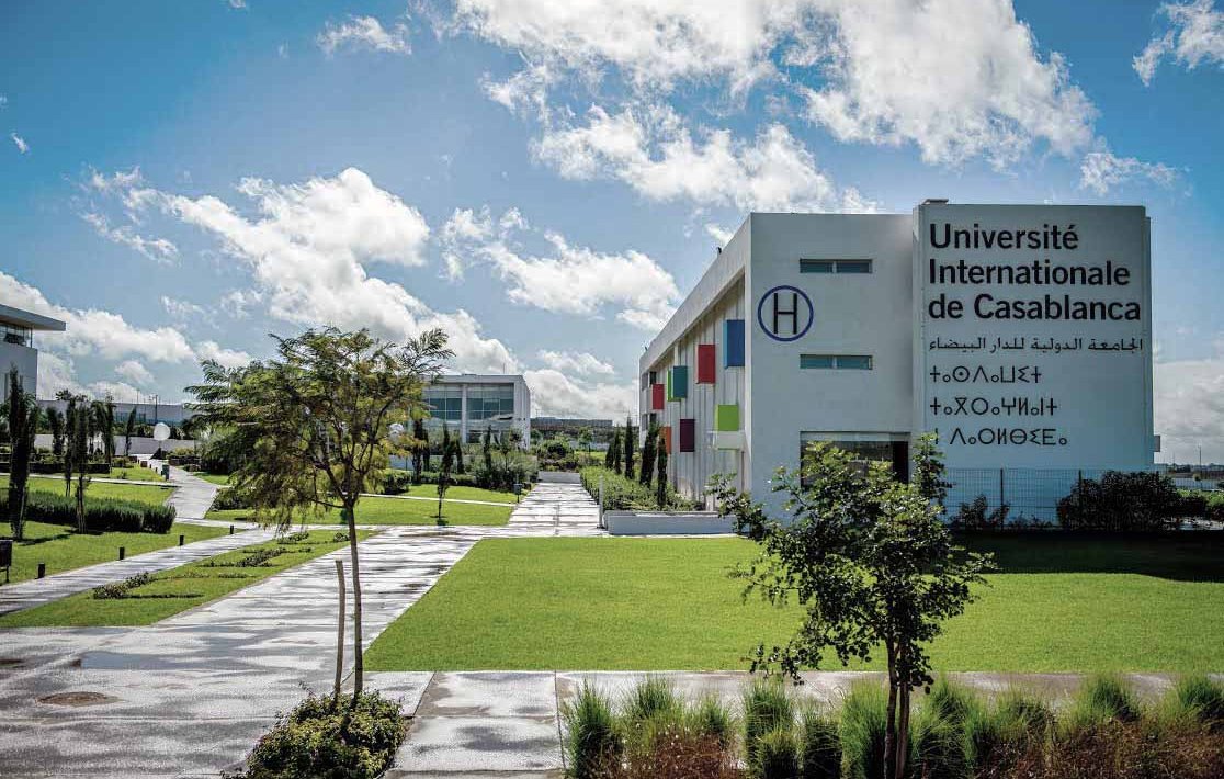 L'image montre l'Université Internationale de Casablanca. On peut voir des bâtiments modernes, avec des façades blanches et colorées. L'environnement est entouré de verdure, incluant des arbres et des espaces verts bien entretenus. Le ciel est bleu avec quelques nuages, et le sol semble humide, suggérant qu'il a peut-être récemment plu. L'ensemble dég dég dég dég dég dég dég dég dég dég dég dég dég dég dég dég dég dég dég dég dég dég dég dég dég dég deg d'une atmosphère accueillante et dynamique.