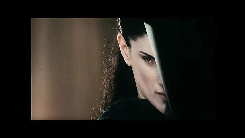 L'image montre une femme avec une expression intense et déterminée. Elle est à moitié cachée derrière un objet, ce qui accentue son regard perçant. Ses cheveux sont longs et foncés, et son attitude suggère qu'elle est dans une situation de confrontation ou de défense. L'éclairage met en avant son visage et crée une ambiance mystérieuse.