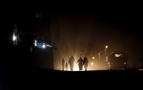 L'image montre un groupe de personnes marchant sur une route sombre, illuminée par des lumières vives au loin. Les silhouettes des individus sont visibles, créant une ambiance mystérieuse. En arrière-plan, on aperçoit des bâtiments et d'autres sources de lumière, suggérant un environnement urbain. L'atmosphère est à la fois sombre et intrigante, mettant en avant le contraste entre l'ombre et la lumière.