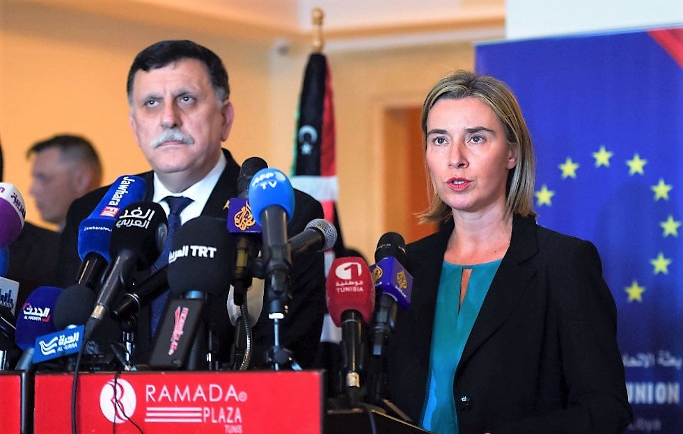 L'image montre deux personnes debout derrière un podium avec plusieurs microphones. La femme, à droite, porte une veste noire et une blouse turquoise, et semble s'exprimer devant les médias. L'homme, à gauche, est également en costume et semble écouter. En arrière-plan, on peut voir des drapeaux et un affichage qui indique une conférence ou un événement officiel. L'ambiance semble être celle d'une conférence de presse.