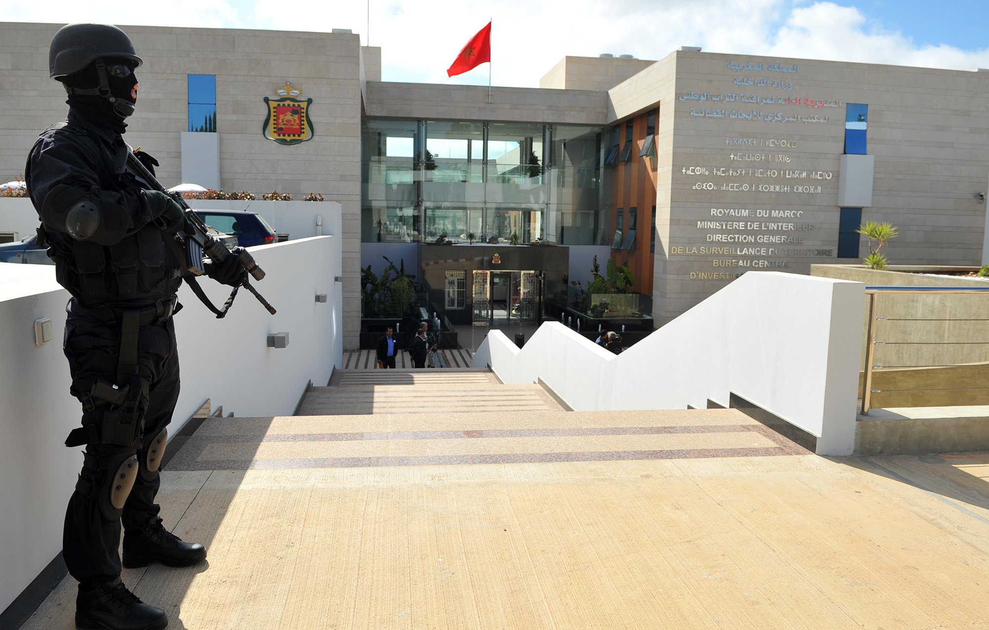 L'image montre un bâtiment moderne et officiel, probablement un ministère ou une institution gouvernementale, orné d'un drapeau marocain. À l'avant, un soldat en tenue noire, portant un masque et une arme, se tient en position de garde sur des escaliers menant à l'entrée du bâtiment. Des inscriptions en français et en arabe sont visibles sur le mur à côté de l'entrée, indiquant le nom de l'institution. L'environnement semble sécurisé et bien entretenu, avec quelques plantes décoratives.