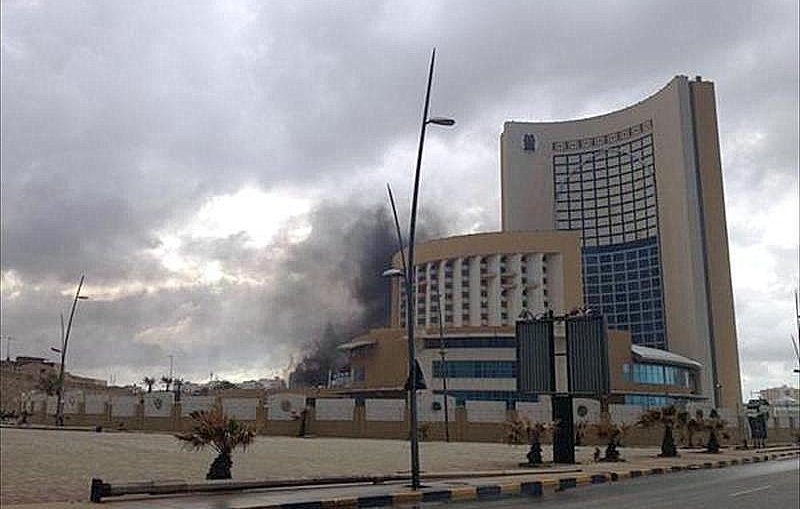 L'image montre un bâtiment moderne de grande taille, probablement un hôtel ou un immeuble de bureaux, situé dans une zone urbaine. À l'arrière-plan, on peut voir de la fumée noire s'élever dans le ciel, ce qui suggère qu'il y a eu un incendie ou une explosion. Le temps semble nuageux, ajoutant une atmosphère sombre à la scène. Des lampadaires bordent la route, et il y a des espaces verts et des plantations autour du bâtiment.