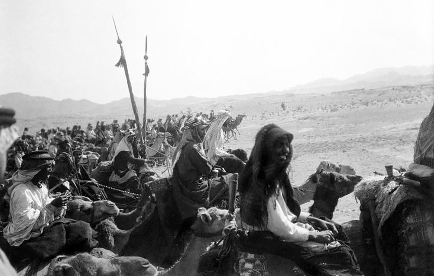 L'image montre un groupe de personnes montées sur des chameaux dans un paysage désertique. On peut voir des femmes et des hommes, certains portant des vêtements traditionnels. Au fond, des montagnes se dressent, et le ciel semble clair. Les personnes semblent participant à une sorte de caravane ou de procession, renforçant l'ambiance historique et culturelle de la scène.