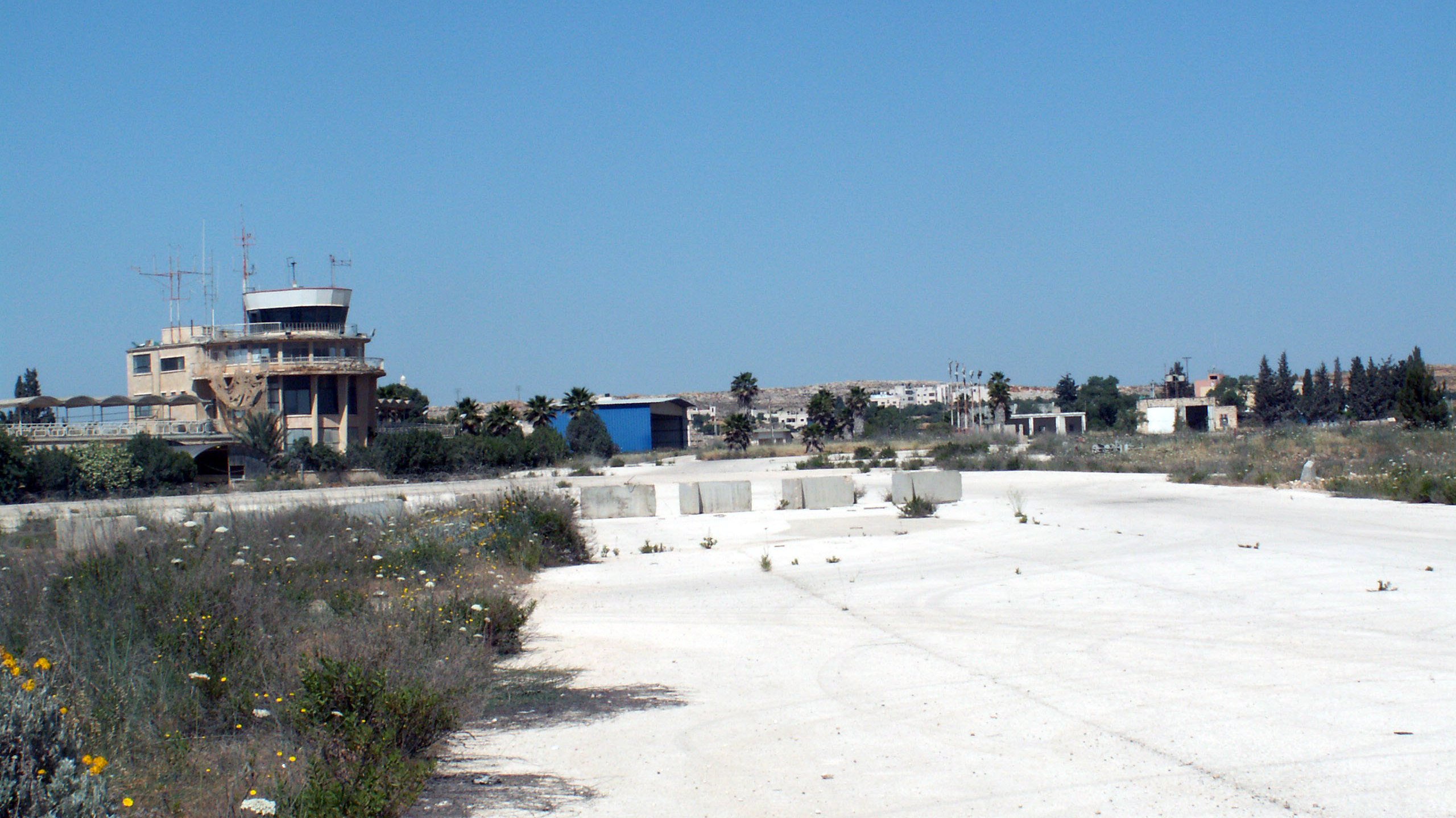 L'image montre un paysage de terrain vague, probablement une ancienne installation ou un aéroport désaffecté. Au loin, on peut voir un bâtiment de type tour de contrôle, avec des antennes sur le toit. Le sol est principalement fait de béton, avec des herbes et des fleurs sauvages qui poussent entre les fissures. Le ciel est dégagé et d'un bleu intense, et en arrière-plan, d'autres structures peuvent être aperçues, suggérant un site qui n'est plus en activité. L'atmosphère semble calme et quelque peu abandonnée.
