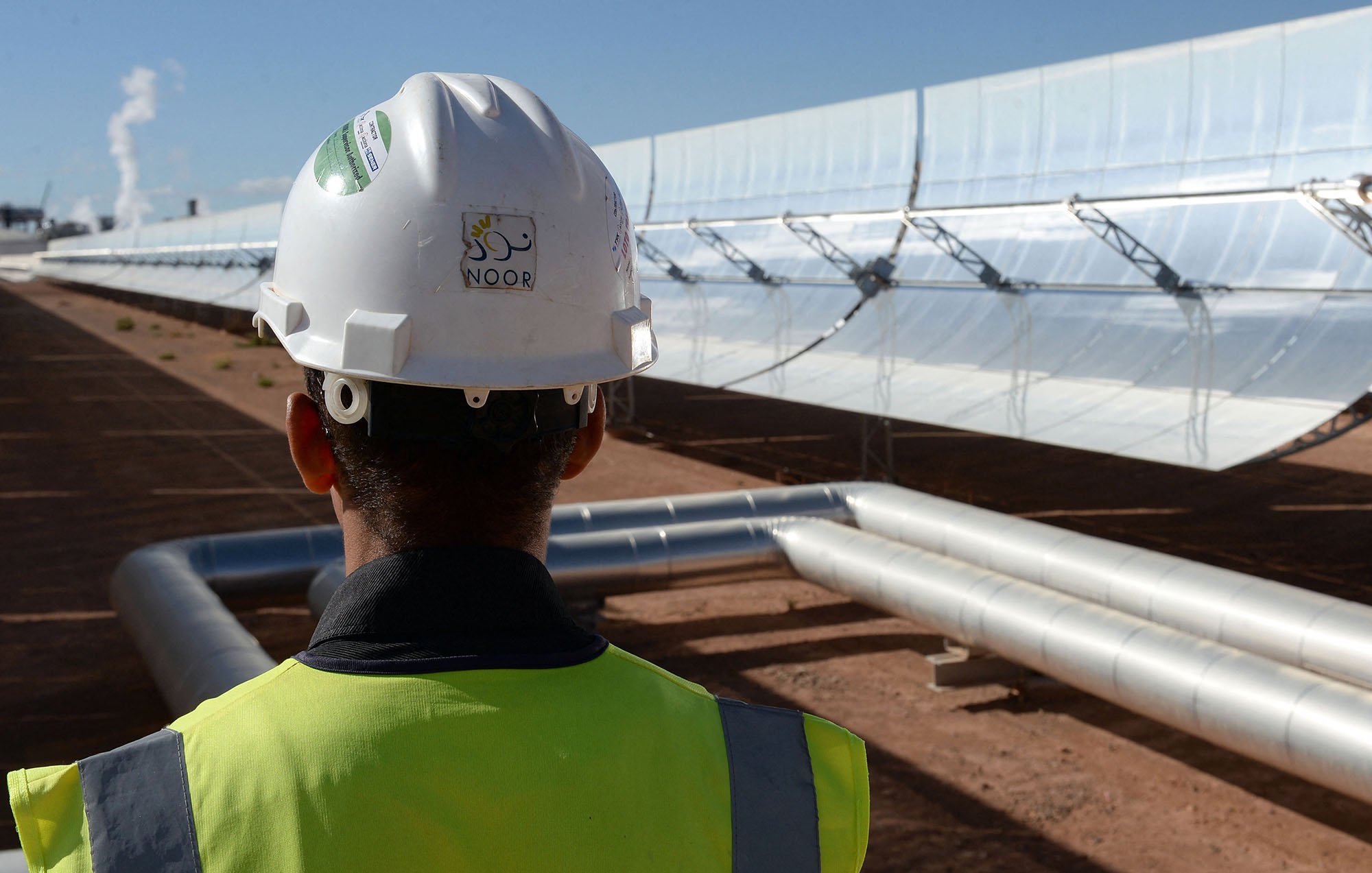 L'image montre un homme portant un casque de sécurité et un gilet de chantier, tourné vers un champ de miroirs solaires. Les miroirs sont orientés vers le soleil et semblent faire partie d'une installation de production d'énergie solaire. À l'arrière-plan, on peut apercevoir des tuyaux métalliques qui transportent probablement un fluide caloporteur. L'environnement est désertique, avec un ciel bleu dégagé qui suggère des conditions solaires idéales.