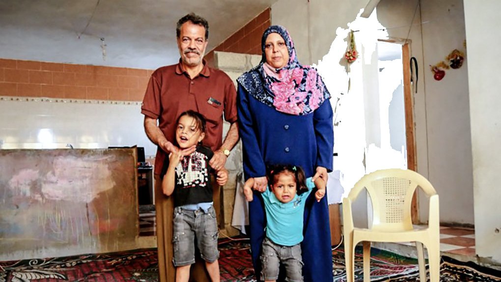 L'image montre une famille pose ensemble dans une pièce chaleureuse. Un homme et une femme, avec deux enfants, se tiennent debout sur un tapis. La femme porte un hijab et une longue robe, tandis que les enfants semblent joyeux. En arrière-plan, on peut voir des murs et une cuisine simple. L'atmosphère semble conviviale et familiale.