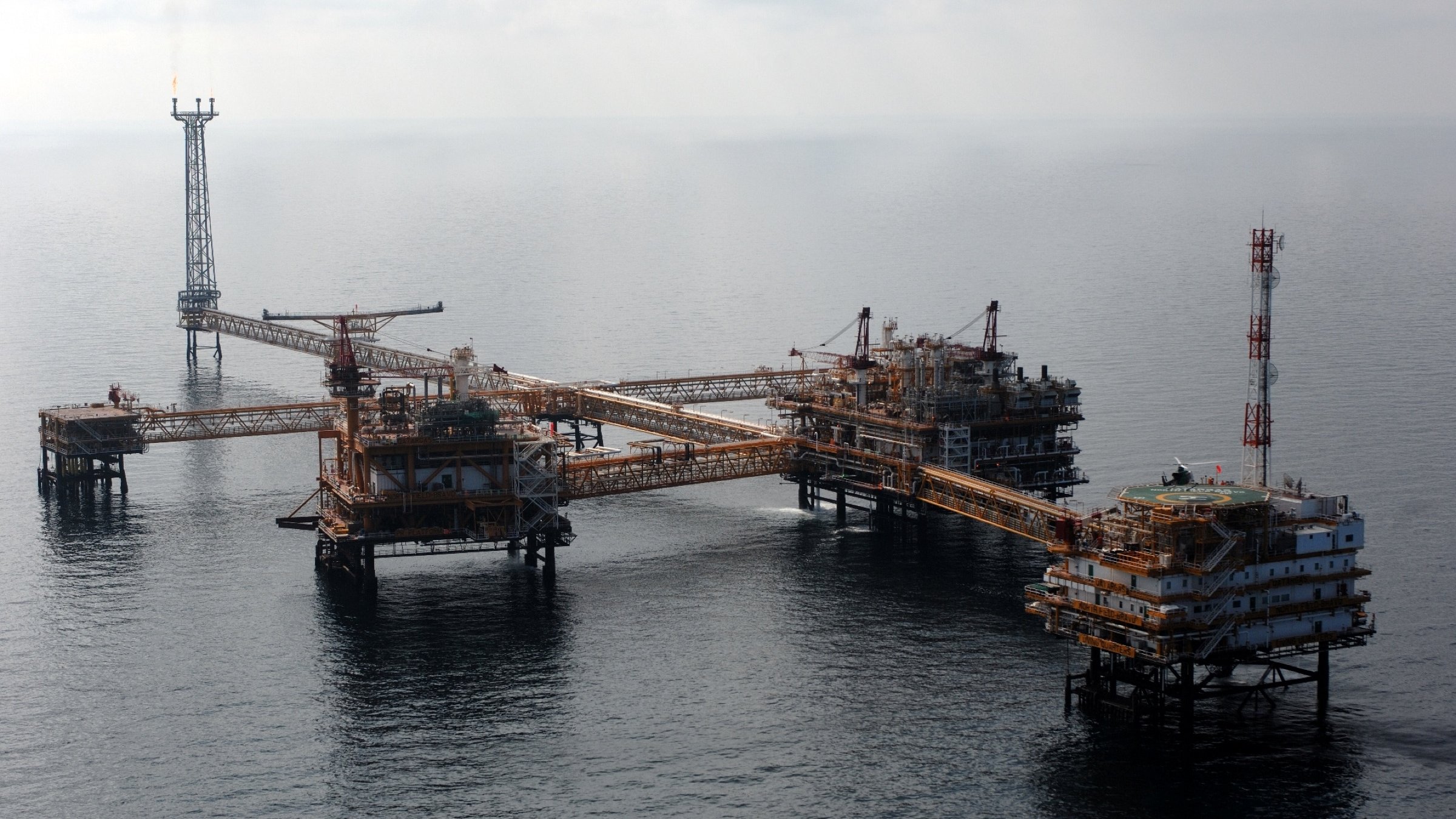 L'image montre une plateforme pétrolière en mer. On peut voir plusieurs structures métalliques assemblées, avec des passerelles reliant les différentes sections. Il y a des installations pour l'extraction du pétrole, ainsi que des antennes et des équipements de communication. La mer environnante est calme, et le ciel est partiellement nuageux. Cette scène évoque l'industrie pétrolière et offshore, avec une infrastructure conçue pour exploiter les ressources sous-marines.