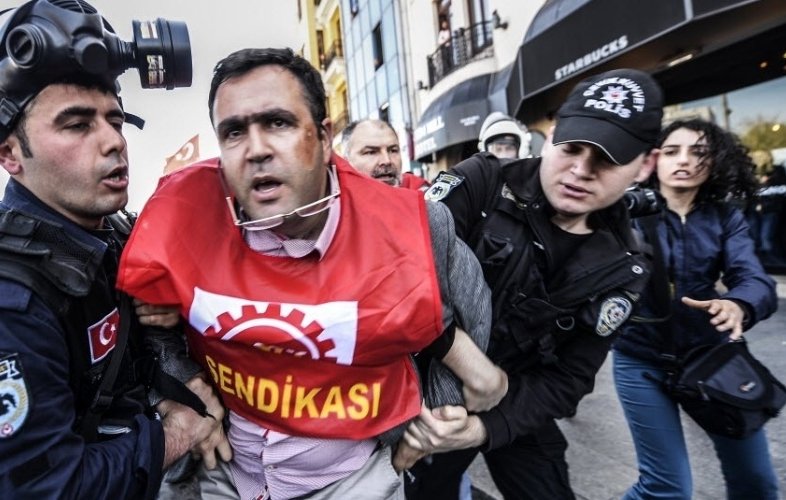 L'image montre une scène de répression où des policiers en tenue d'intervention arrêtent un homme qui porte un t-shirt rouge avec le mot "SENDIKASI". L'individu semble être en train d'être mené à l'écart, visiblement en désaccord avec son arrestation. Les policiers sont équipés de casques et semblent concentrés sur leur opération. L'environnement est urbain, et on peut voir un café Starbucks en arrière-plan, ce qui suggère que cette scène se déroule dans une zone animée de la ville.