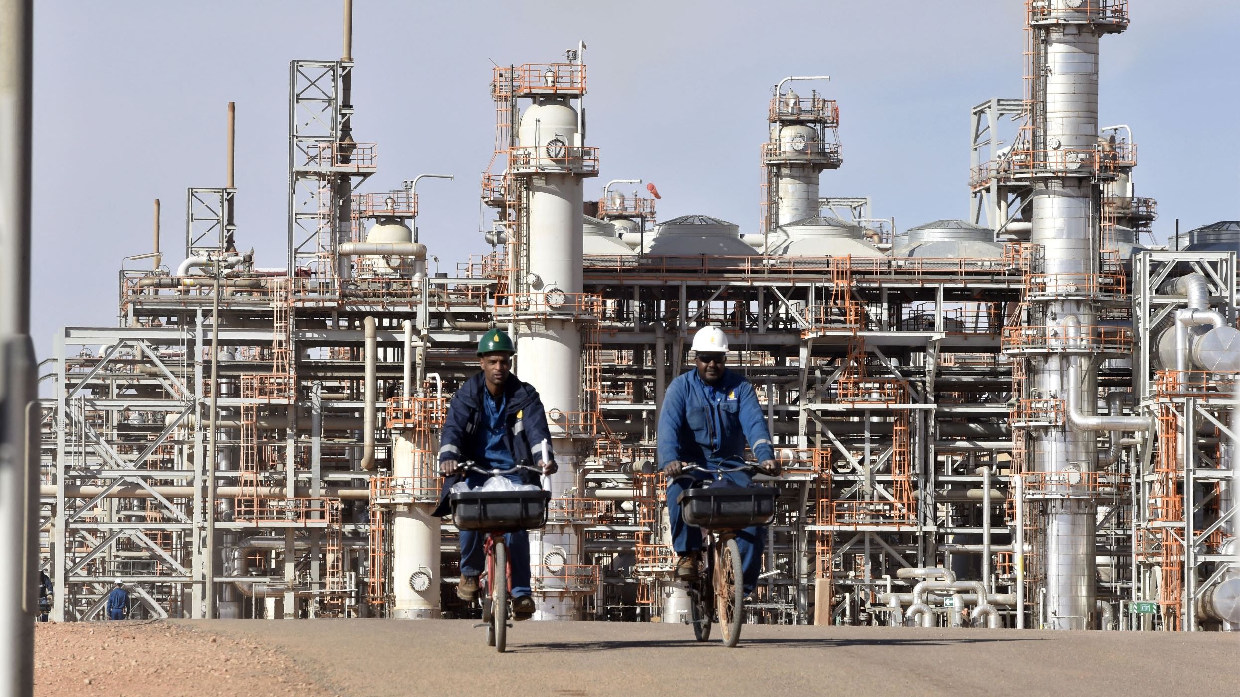 L'image montre deux hommes à vélo, portant des vêtements de travail et des casques, qui circulent sur une route. En arrière-plan, on peut voir une installation industrielle avec de grandes structures métalliques et des pipelines, typiques d'une raffinerie ou d'un complexe pétrochimique. Le ciel est clair et les installations semblent actives, témoignant d'un environnement de travail industriel.