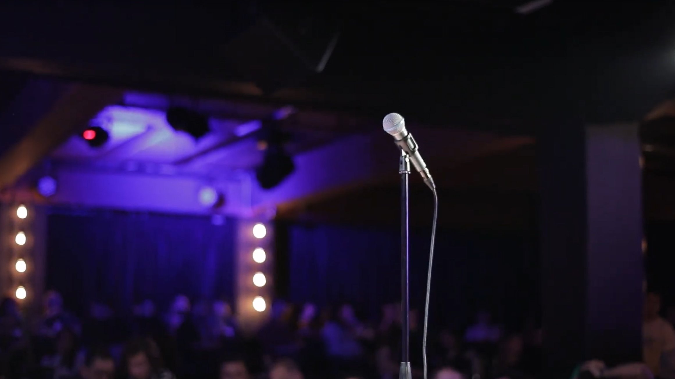 L'image montre un microphone sur un pied, au premier plan, sur une scène sombre. À l'arrière-plan, on aperçoit une audience floue et des lumières tamisées qui créent une ambiance de spectacle.