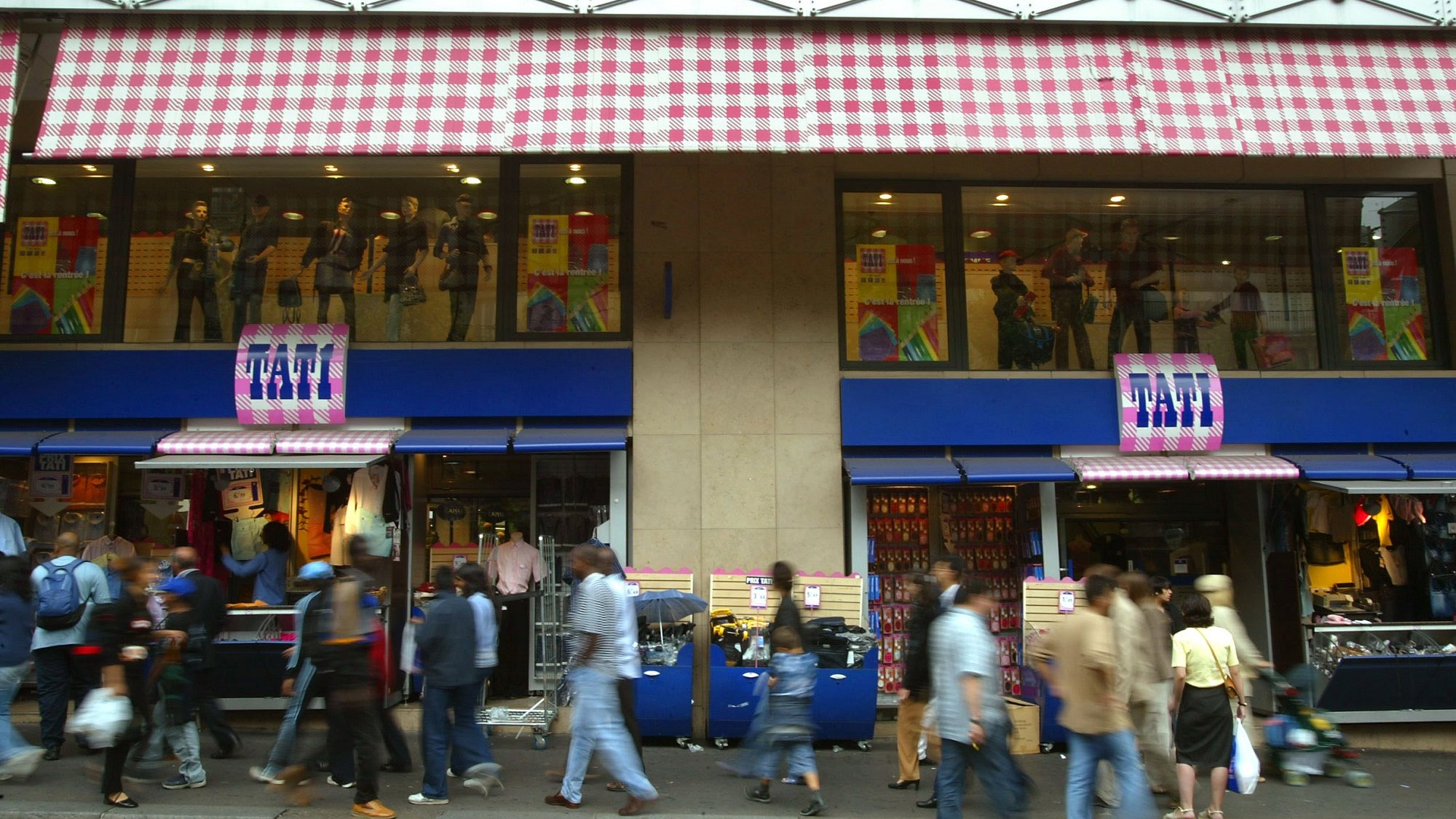 L'image montre une rue animée avec des piétons marchant devant un magasin. Le magasin est identifié par un enseigne "FATI" et présente des vitrines avec des mannequins habillés. Les façades supérieures du magasin ont des stores à carreaux. On peut voir une multitude de personnes se déplacer sur le trottoir, créant une atmosphère dynamique et vivante.