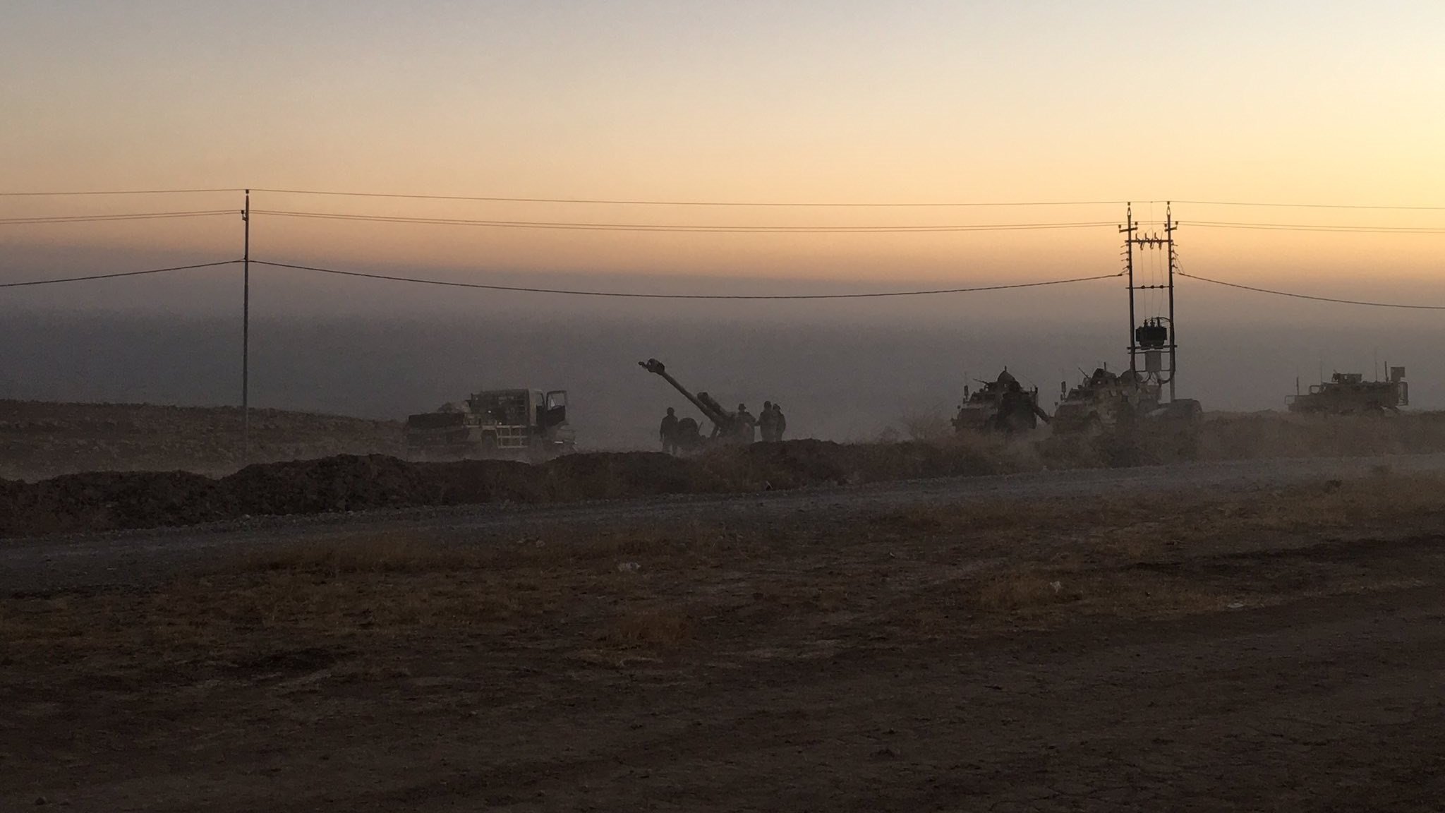 L'image montre un paysage de chantier au crépuscule, avec un ciel teinté de couleurs douces allant du jaune à l'orange. On aperçoit des machines de construction et des travailleurs en silhouettes, entourés de poussière. À l'arrière-plan, on distingue des poteaux électriques. Le sol semble sec et rocailleux, suggérant une zone de travaux en cours. L'atmosphère est calme, mais il y a une impression d'activité et d'effort en cours.